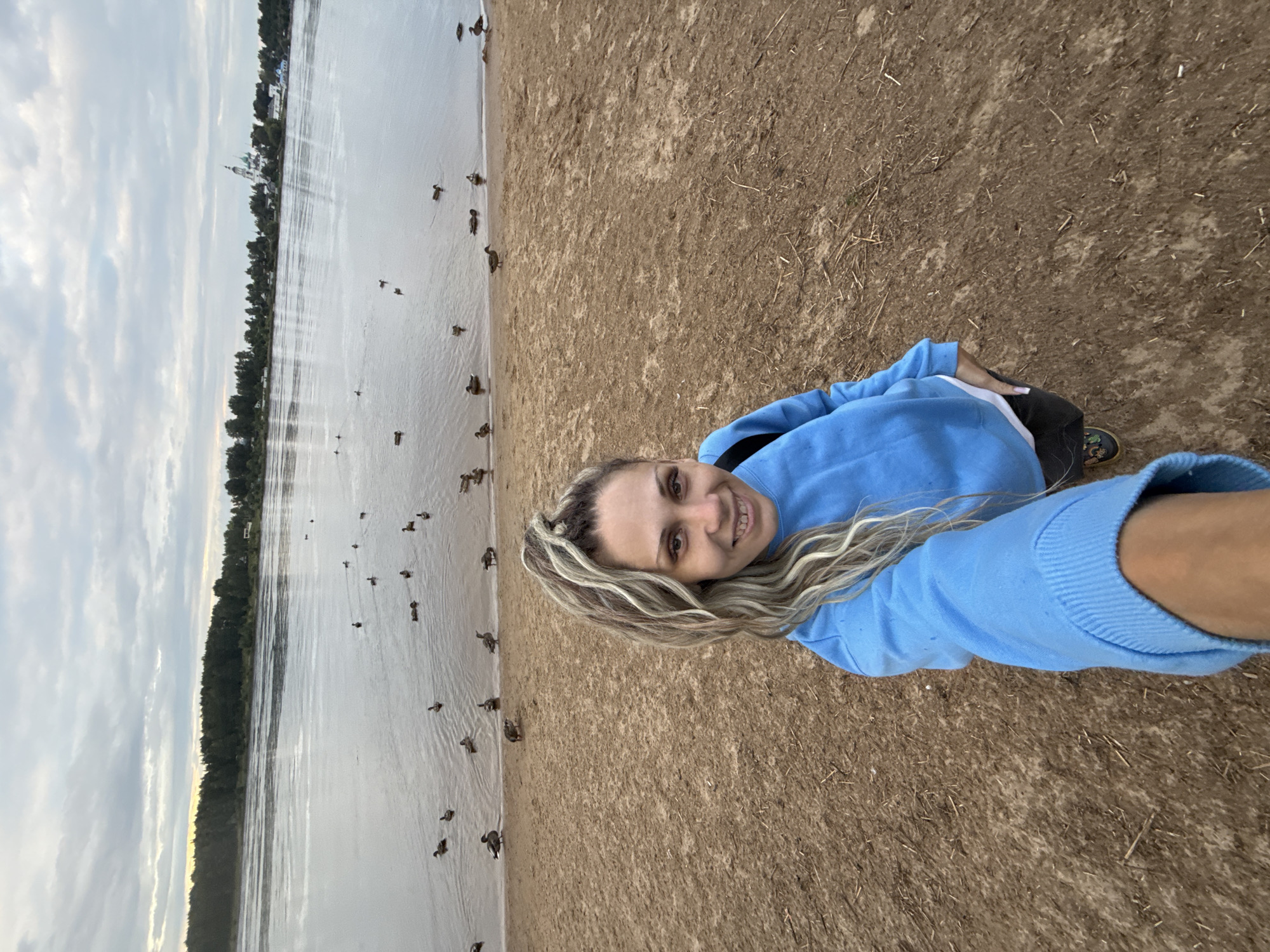 A woman with blonde hair tied in a high ponytail takes a selfie on a sandy beach. She is wearing a bright blue sweatshirt and black pants. Behind her, a wide river stretches towards a treeline, with several ducks swimming in the water. In the distance, a white church with green domes is visible. The sky is overcast with scattered clouds. The scene is set in Rusanovo, Russia.