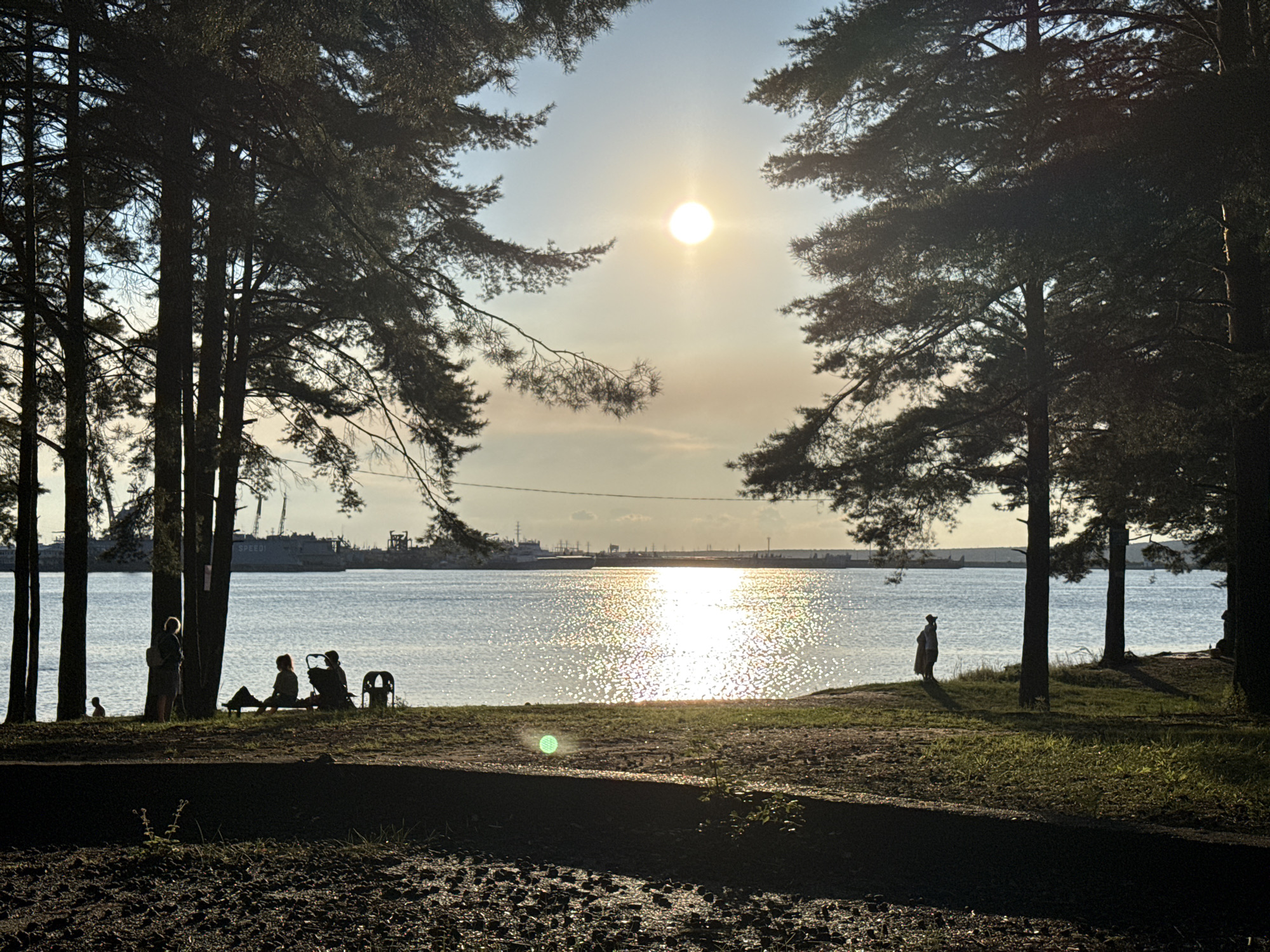 The image depicts a sunlit body of water with several figures silhouetted against the bright sky and reflecting sunlight. The scene appears to be a park or shoreline area with mature trees framing the view of the water. In the foreground, dark soil and grass are visible.
On the left side of the image, near the water's edge, a person stands with their back to the camera, wearing a backpack. To their right, a group of people is seated on a bench or low structure, with what appears to be a stroller or carrier nearby. Further to the right, closer to the water, another solitary figure stands on a grassy embankment.
The water is calm and reflects the bright sun, creating a shimmering effect. In the distance, across the water, a line of industrial structures or ships is visible along the horizon, with cranes and masts silhouetted against the hazy sky. The sun is positioned high in the sky, creating a warm, late afternoon or early evening light. The overall atmosphere is serene and peaceful.
While the image does not explicitly state the location within Chaikovsky, Russia, the presence of a large body of water, trees, and industrial elements in the distance is consistent with the landscape of the city, particularly areas along the Kama River.