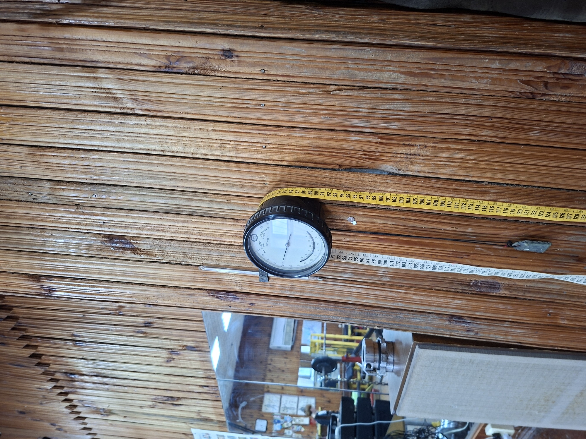 The image depicts a close-up of a wooden wall, likely in a gym or workshop setting. A circular gauge, possibly a barometer, is mounted on the wall, with a yellow measuring tape hanging vertically beside it. The tape displays numerical markings from 84 to 131. Reflections in a mirror to the left suggest the presence of exercise equipment, including weight machines and barbells. The overall aesthetic is rustic, with the textured wooden surface dominating the frame. The location is specified as a gym in Kazan, Russia.