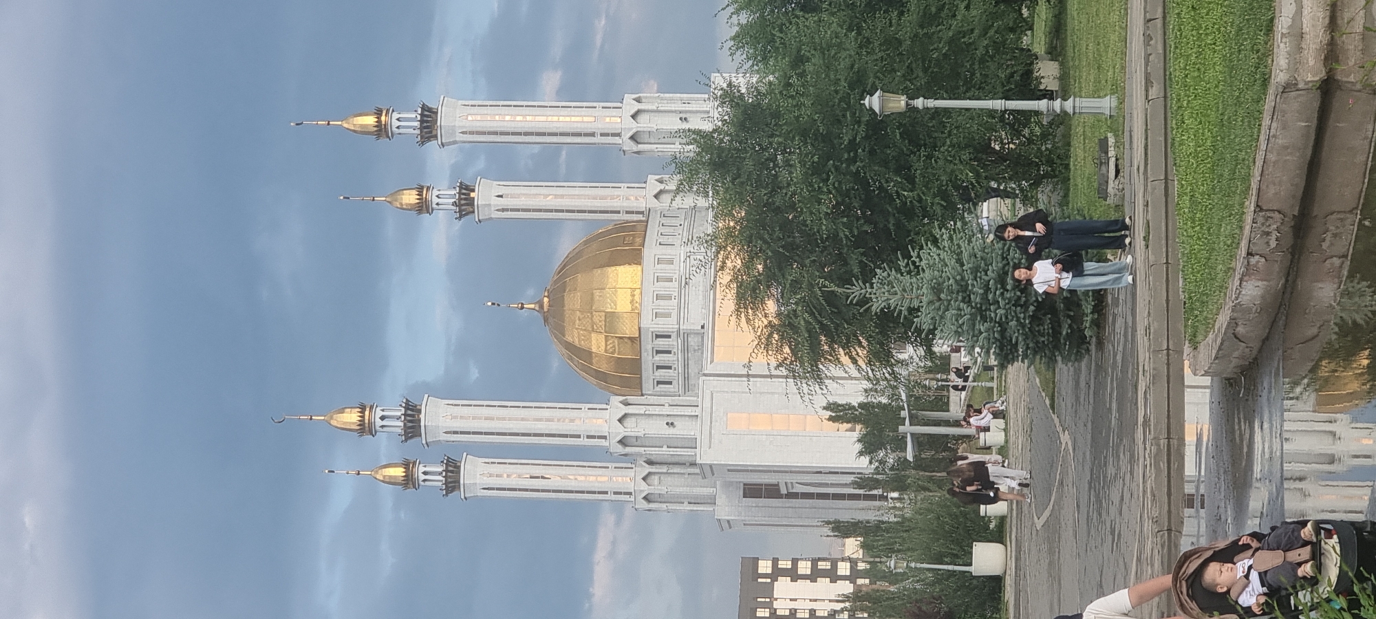 The image shows the Nur-Gasyr Mosque, a landmark in Aktobe, Kazakhstan. The mosque features a large golden dome and four tall minarets, characteristic of Islamic architecture. The building is predominantly white with gold accents. In the foreground, there is a paved area with several people, including two young girls standing and looking forward, and a baby in a stroller. To the left, a modern apartment building is visible. The scene is set within a park-like environment with trees and lampposts, under a cloudy sky. The ground is wet in places, possibly from recent rain, reflecting the surroundings.
