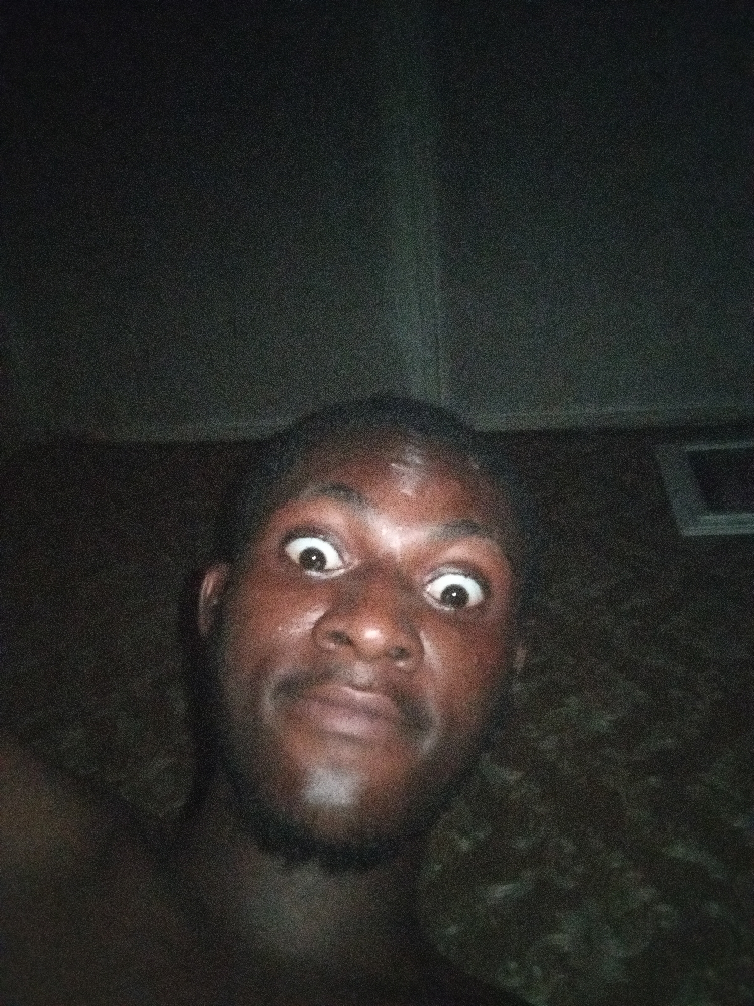 A close-up selfie of a dark-skinned man with wide eyes. He has short, dark hair and a beard. The background is dark. A window-like shape is visible in the upper right. The location is Unknown, Unknown.