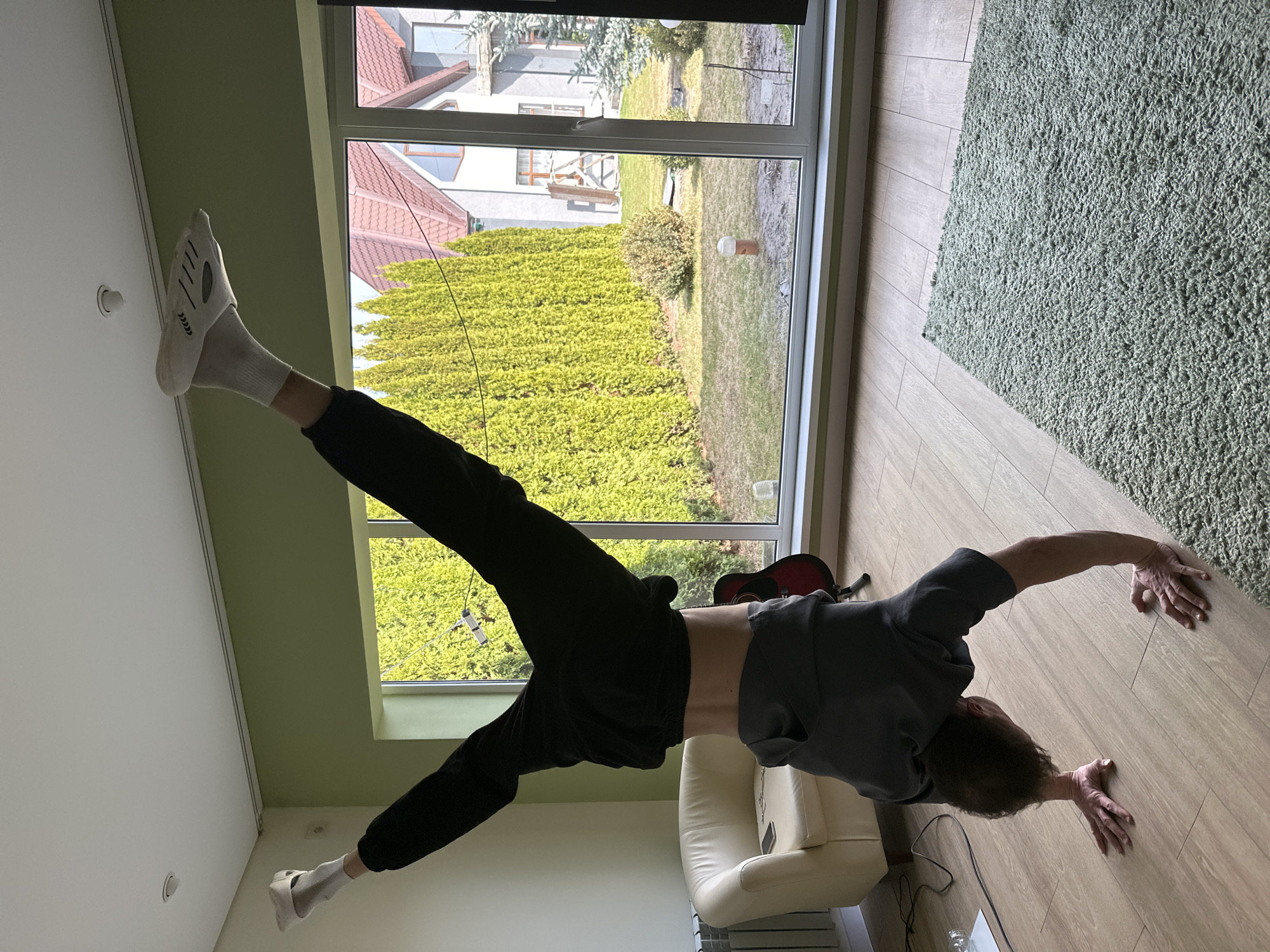 A person is performing a handstand with legs split near a window in an indoor setting. The location of Kursk, Russia cannot be determined from the image. The person is wearing dark-colored sweatpants, socks, and a gray shirt. Outside the window, there is a green hedge and a view of neighboring houses and yards. The room has light-colored walls and wooden flooring. A white armchair is visible in the background. A green rug is on the floor.