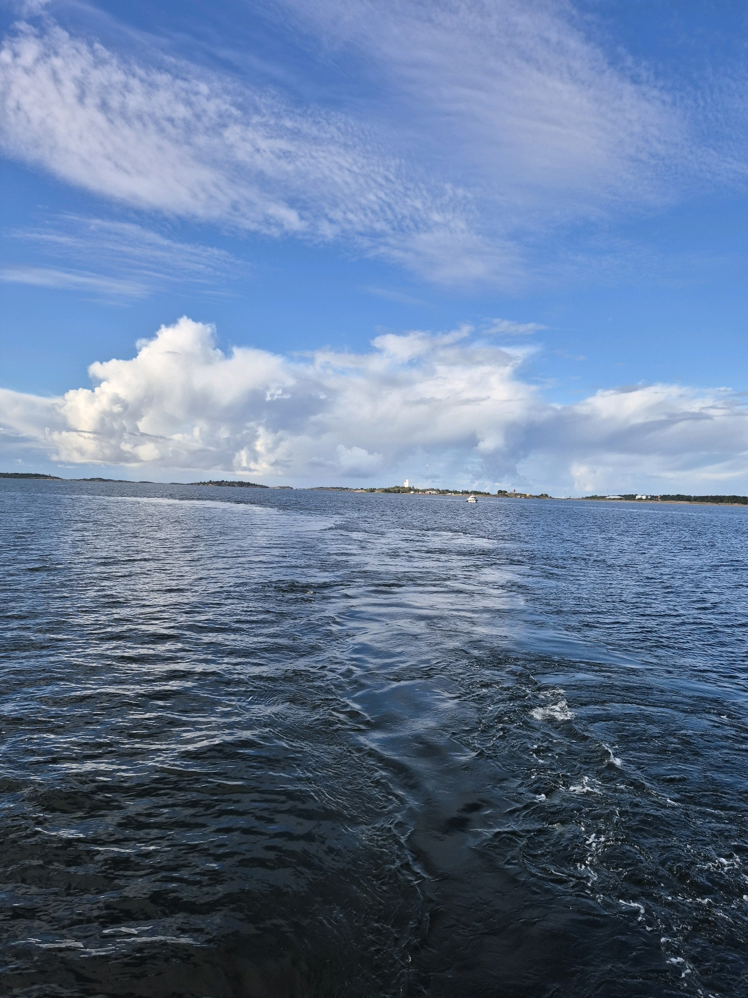 The image depicts a view from a boat on the sea, looking towards the horizon. The water in the foreground is dark blue and choppy, with a visible wake trailing behind the boat. In the distance, a landmass with buildings and trees is visible under a partly cloudy sky. The sky is a vibrant blue with streaks of white clouds. A small white boat is visible on the water in the middle distance. This scene is characteristic of the waters surrounding Hanko, Finland, a coastal city known for its archipelago and maritime activities.
