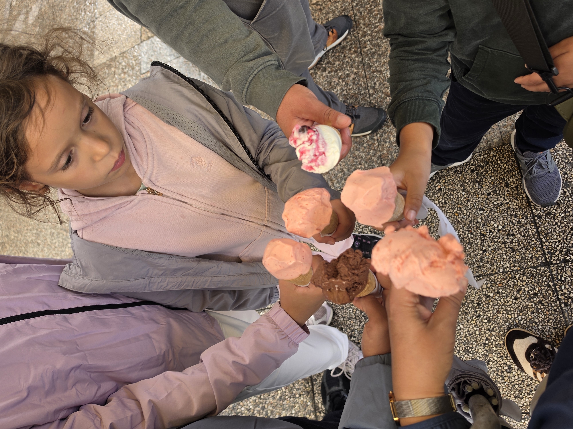 A group of at least six people, including a young girl in the center, are gathered together, holding ice cream cones. The girl, looking down, wears a pink hoodie and a gray jacket. Other individuals are seen from the chest down, wearing casual clothing such as jackets and pants in shades of pink, purple, gray, and green. They are all holding ice cream cones, with visible flavors including pink (likely strawberry or raspberry), chocolate, and a white ice cream with pink swirls. The background appears to be an outdoor setting with a textured ground surface, possibly cobblestone or mosaic paving, indicative of an urban environment like San Giovanni, Italy. The ground is a mix of light and dark speckled aggregate. The people are in close proximity, suggesting a shared activity.