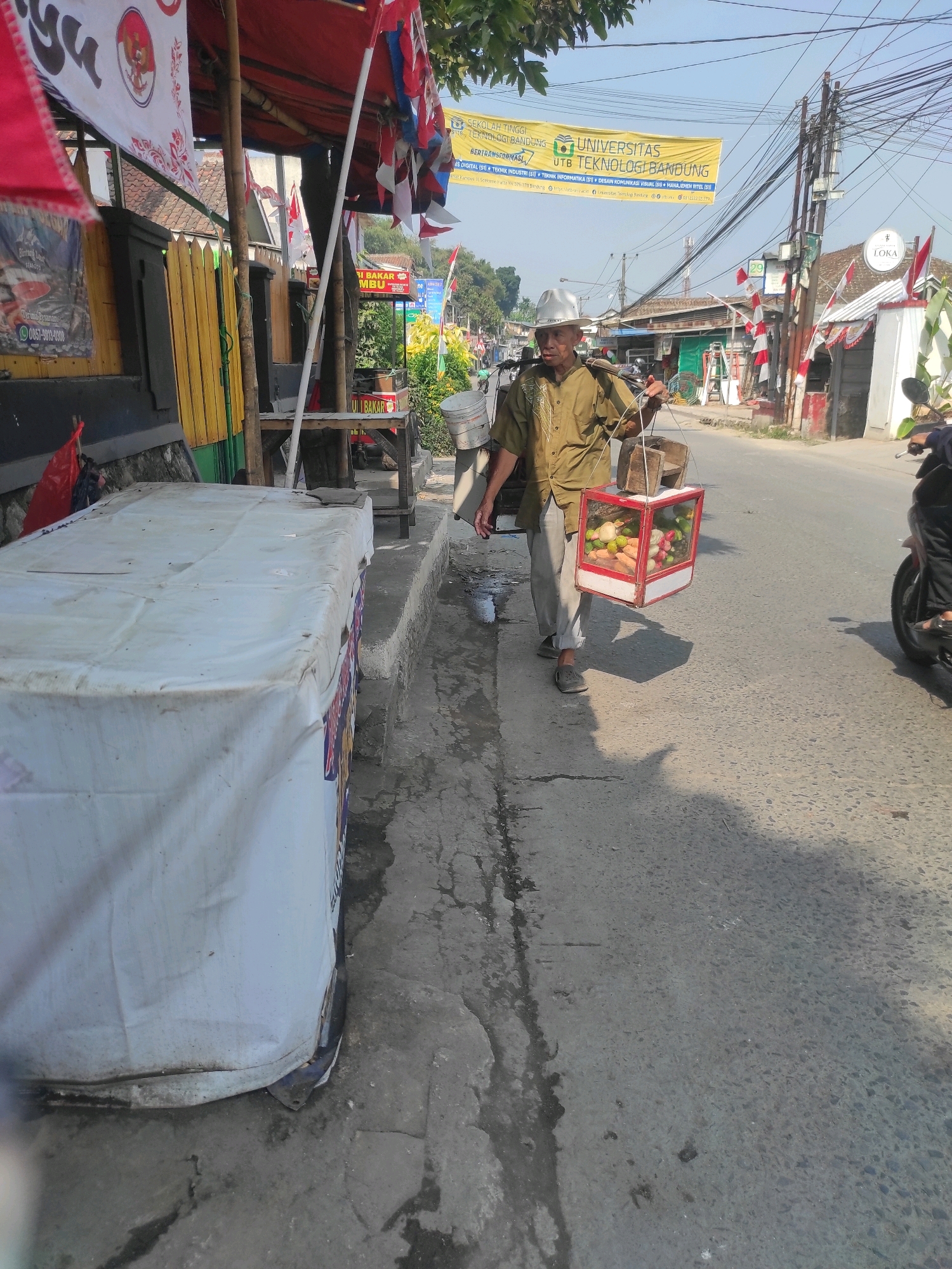 A man is walking on a sidewalk next to a street in Kertamulya, Indonesia. He is carrying a box filled with fruit on a stick over his shoulder. The box is red and white and has glass panels. He is wearing a tan shirt, gray pants, and a white hard hat. The man is walking away from the camera. To the left of the man, there is a white table covered with a tarp. Behind the table, there is a building with yellow and red accents. In the background, there are trees and buildings, and a banner with text in Indonesian that says "SEKOLAH TINGGI TEKNOLOGI BANDUNG UNIVERSITAS UTB TEKNOLOGI BANDUNG". The street is paved and there are vehicles and people in the distance. The sky is clear and the sun is shining.