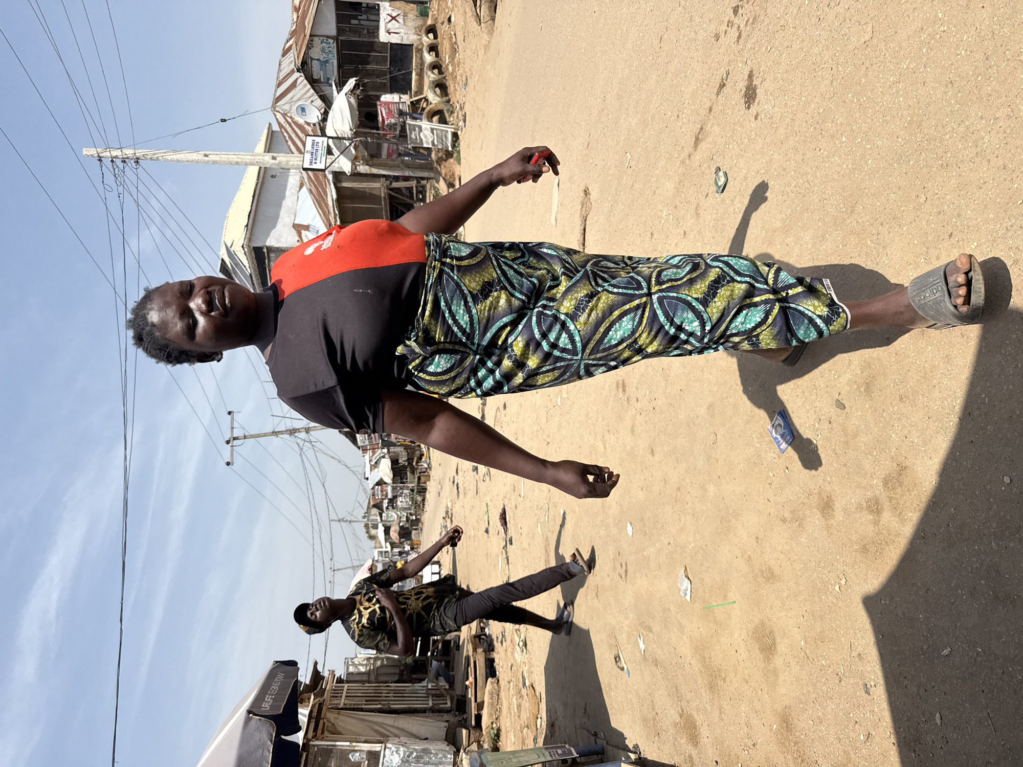 In a sun-drenched street in Abuja, Nigeria, a woman with dark skin and short, dark hair walks towards the camera. She is wearing a two-toned shirt, black on the left and red on the right, and a long, patterned skirt with blue, green, and yellow designs. Her right hand is extended slightly forward, holding a small, red object. In the background, a young man with dark skin wears a patterned black shirt with gold accents and dark pants, walking away from the camera with his head tilted upwards and his eyes closed. He is wearing black baseball cap and gray sandals. The street is unpaved and dusty, with scattered debris, including plastic wrappers and a green straw. Several buildings with corrugated metal roofs line the street, some with open storefronts. Utility poles with numerous wires are visible against a pale blue sky with wispy clouds. A sign for "DREAM LODGE & SUITES LTD" is mounted on a pole. The overall scene depicts a busy, open-air market or residential area on a bright day.
