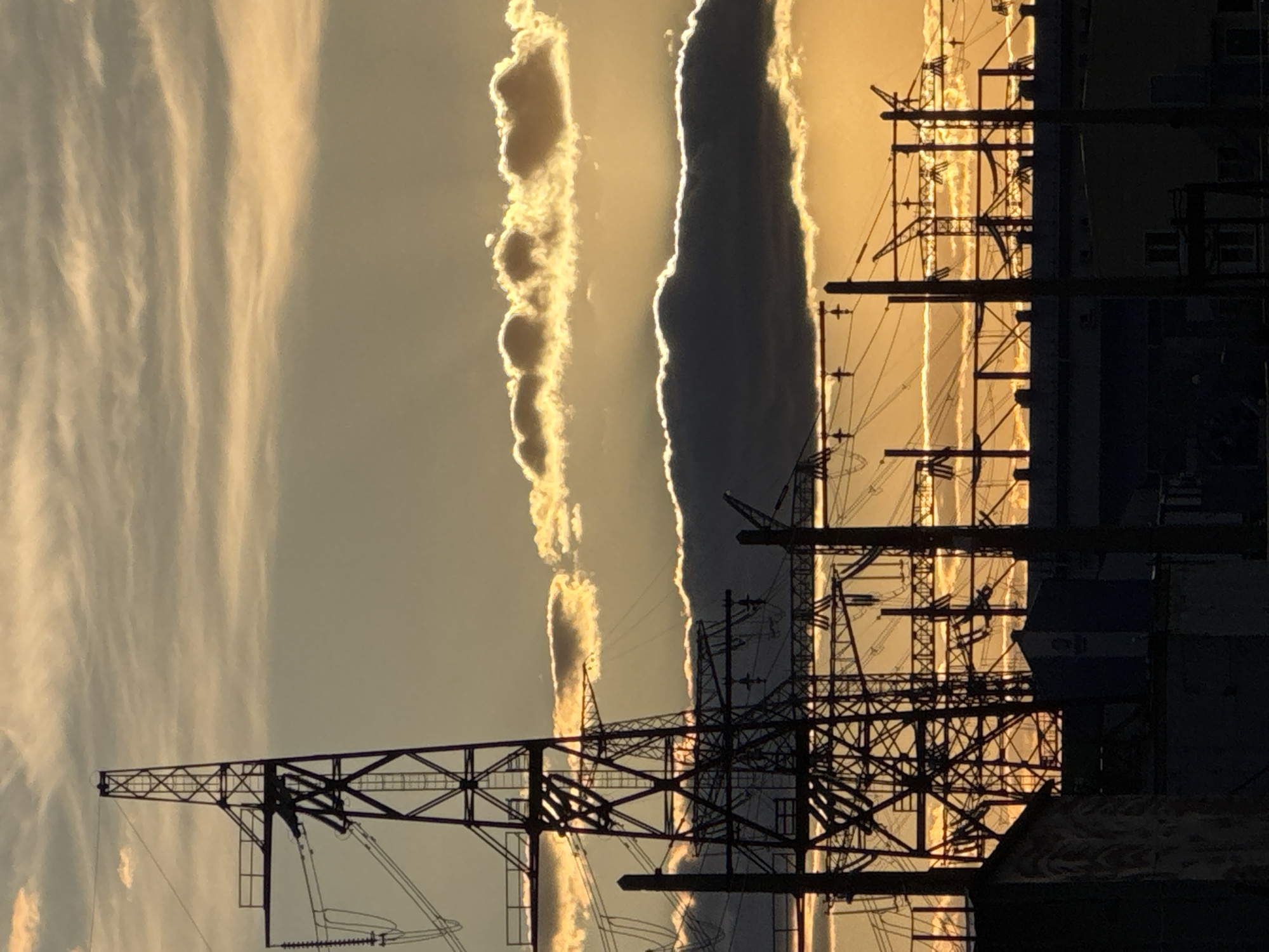 The image captures a view of power lines and support structures silhouetted against a dynamic sky at sunset or sunrise. The setting is likely in or around Volzhsky, Russia, given the presence of industrial infrastructure. Two distinct cloud formations are visible: one with layered, wavy edges and another with a dark, more solid appearance. The power lines form an intricate web, obscuring the sky in places. A building or structure is visible on the right edge of the frame, partially obscured by the power grid. There are no people visible in the image.