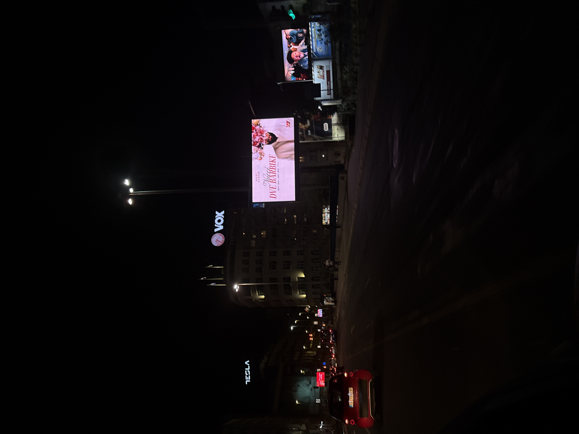 The image depicts a night scene in Belgrade, Serbia, featuring urban elements. A red Fiat 500 is in the foreground, with the license plate "BG-1669 AB" visible. Street lights illuminate the road. Above, a "Tesla" sign is visible on the left. A taller building displays the "VOX" logo higher up. Billboards are present, one promoting the album "DVE BARBIKE" by Naporan dan, while another shows the brand name "MS". The overall atmosphere suggests a busy urban environment at night.