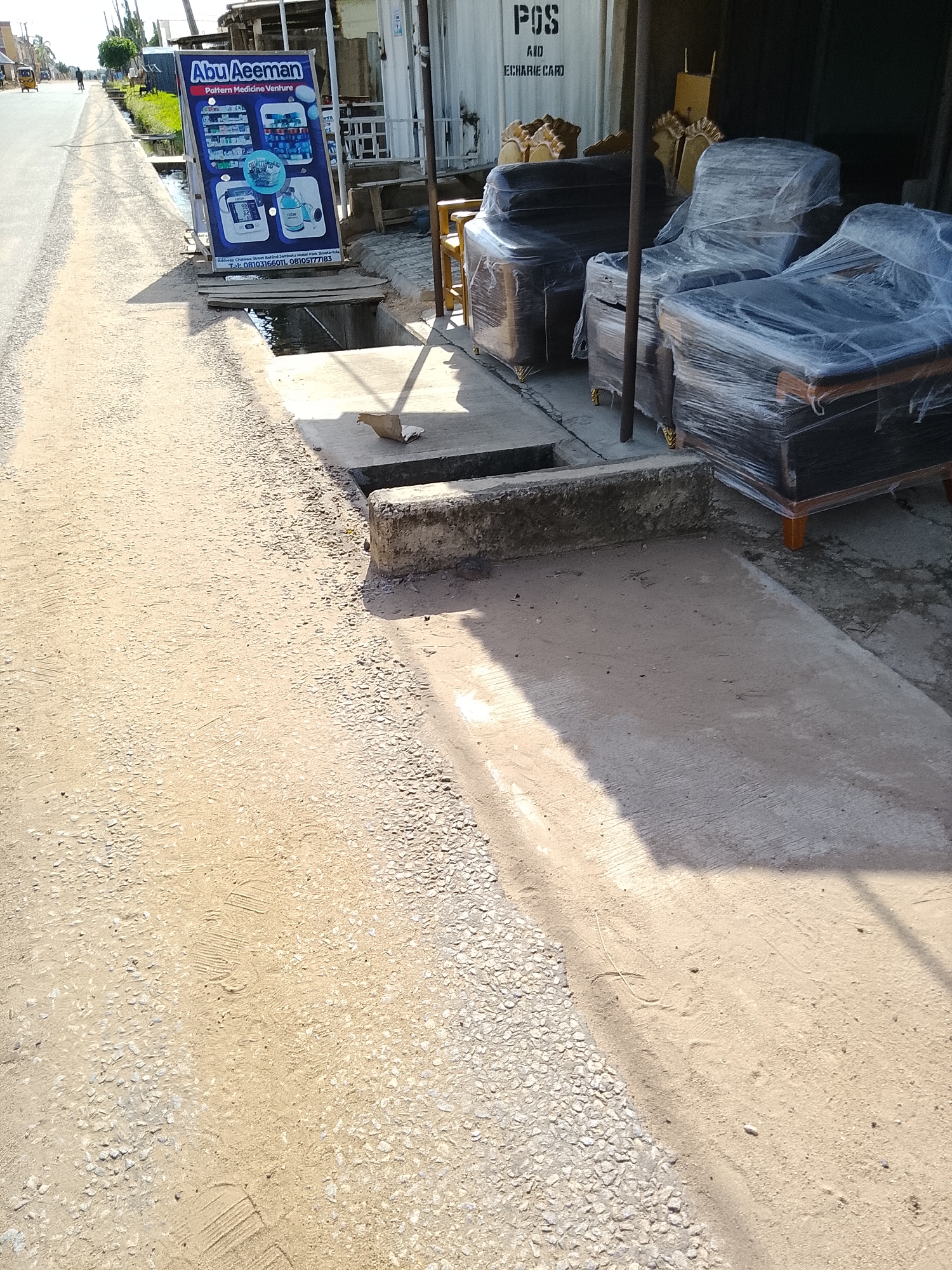 The image captures a street scene in Wauro Leggal, Nigeria. A sign for "Abu Aeeman Pattern Medicine Venture" stands prominently on the left, displaying various medical products and contact information. To the right, furniture wrapped in plastic is displayed outdoors, suggesting a shop or business. The ground is a mixture of dirt, gravel, and a concrete pathway, with a drainage ditch running alongside the road. A few people are visible in the distance on the road, and a tricycle-like vehicle is also present. The environment appears to be a commercial area with a mix of residential and business establishments.