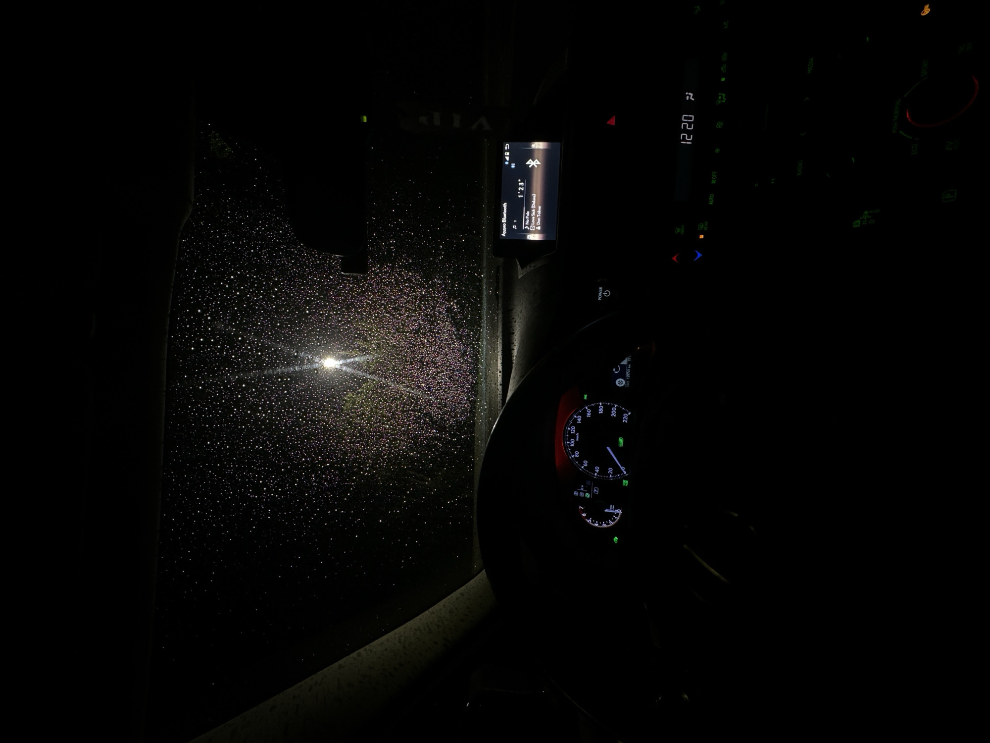 The image depicts the interior of a car at night, during a rainstorm. The windshield is covered in raindrops, reflecting the light from a streetlamp outside. Inside the vehicle, the dashboard is illuminated, displaying the speedometer, the car's audio system, and climate control settings. The audio system indicates a song playing from "Audio Bluetooth" with "1'23"" elapsed, with title "No Pole", artist Dekave, and featuring Don Toliver. The time on the dashboard reads "12:20". The location appears to be within a city, as evidenced by the presence of street lighting.