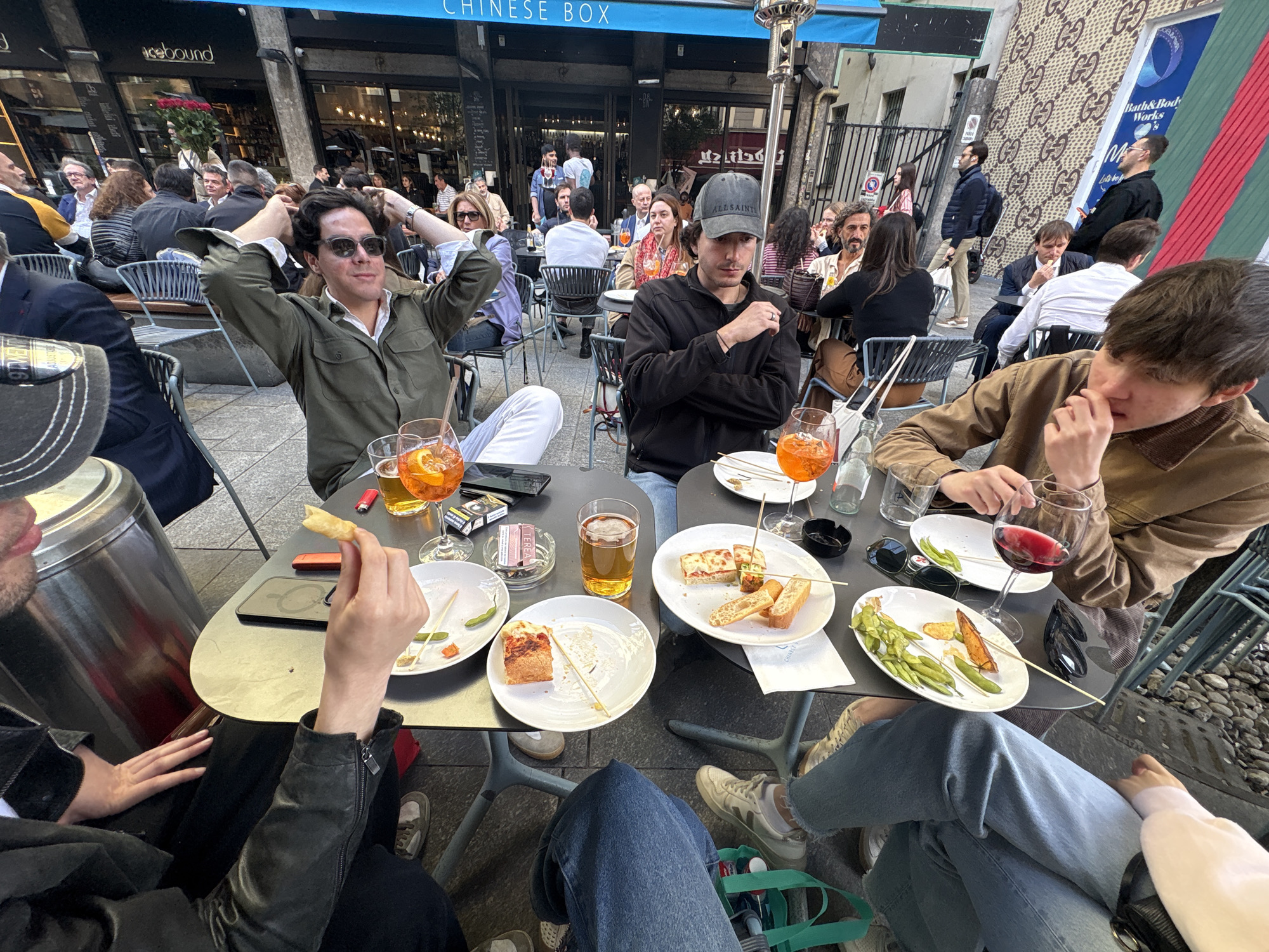 The image depicts a group of people gathered around tables at an outdoor cafe in Milan, Italy. The cafe appears to be located on a busy street. The tables are filled with plates of food, drinks, and personal items. The individuals at the tables are engaged in conversation and enjoying their meals. There is a "Chinese Box" restaurant sign in the background. A Gucci logo wall is visible on the right side of the image, suggesting the location could be in or near the Quadrilatero della Moda, Milan's fashion district. People are walking by in the background.