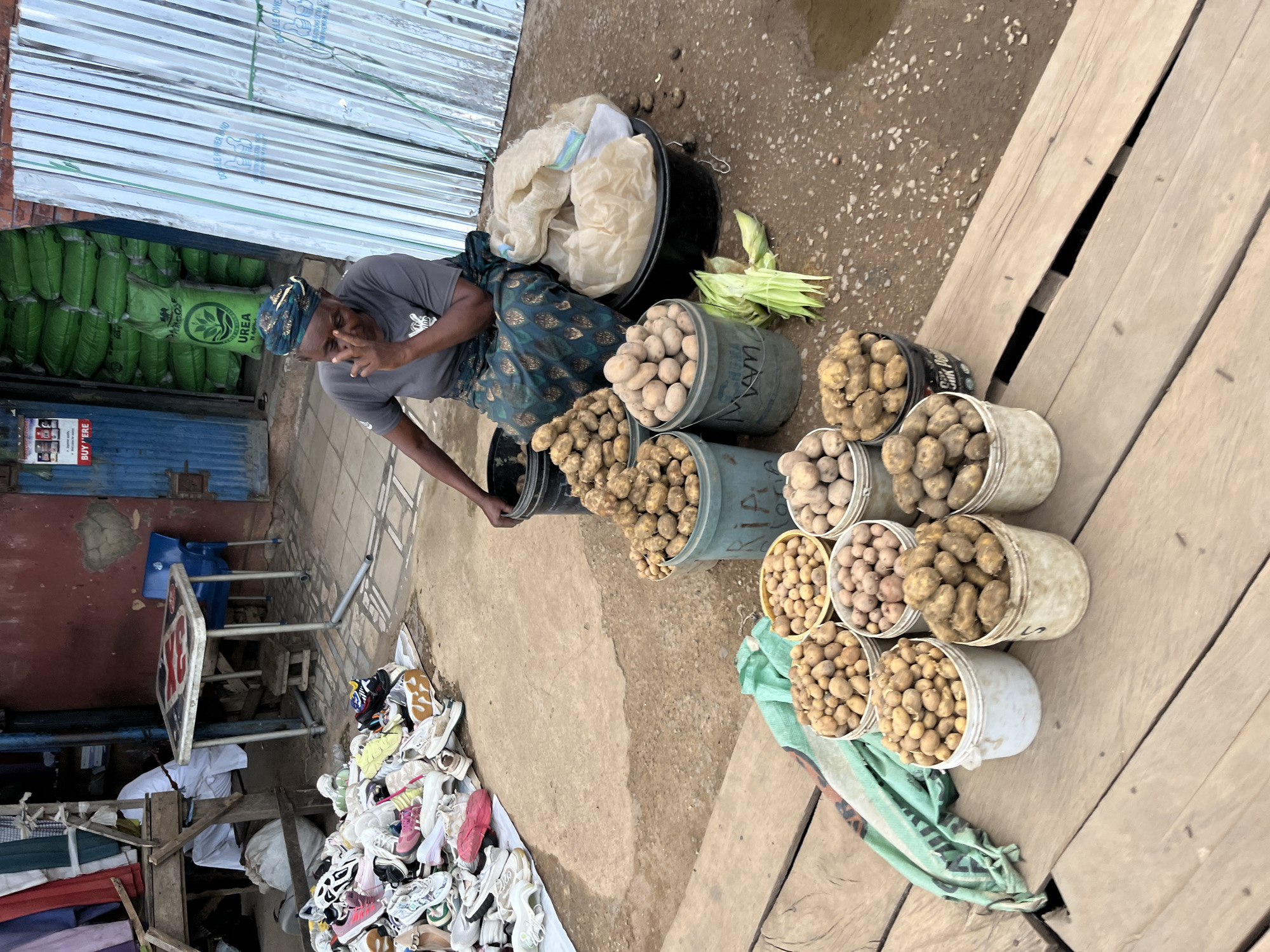 A woman in a headwrap and patterned skirt sits at a market stall in Bukuru, Nigeria. She is wearing a grey t-shirt and her hand is raised to her face, possibly scratching her eye. In front of her, several buckets overflow with potatoes. To her left, a pile of assorted shoes sits on the ground. Behind her, a stack of large green bags labeled "UREA" are visible, along with a corrugated metal structure. The ground is a mixture of dirt and gravel.