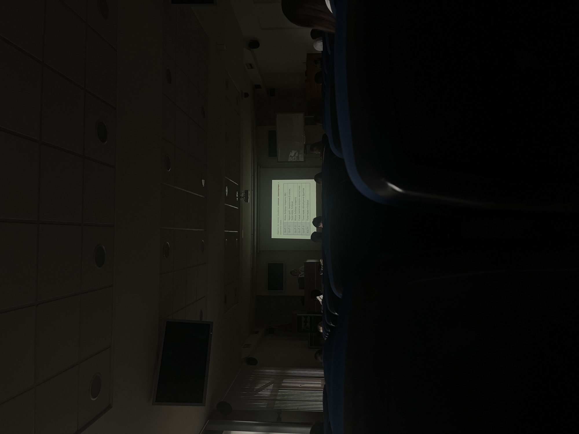 This image shows an indoor lecture or presentation in a room with rows of dark blue seats. A projector screen displays a text list with the following terms: GLUT-1, GLUT-2, GLUT-3, GLUT-4, GLUT-5, SGLT-1, and descriptions related to organs like the brain, kidneys, liver, and intestines. People are seated in the audience, partially obscured by the dark lighting and seat backs. A flat-screen television is mounted on the wall to the left. Blinds are visible behind the screen. The location is described as being in the city of Unknown, Unknown.