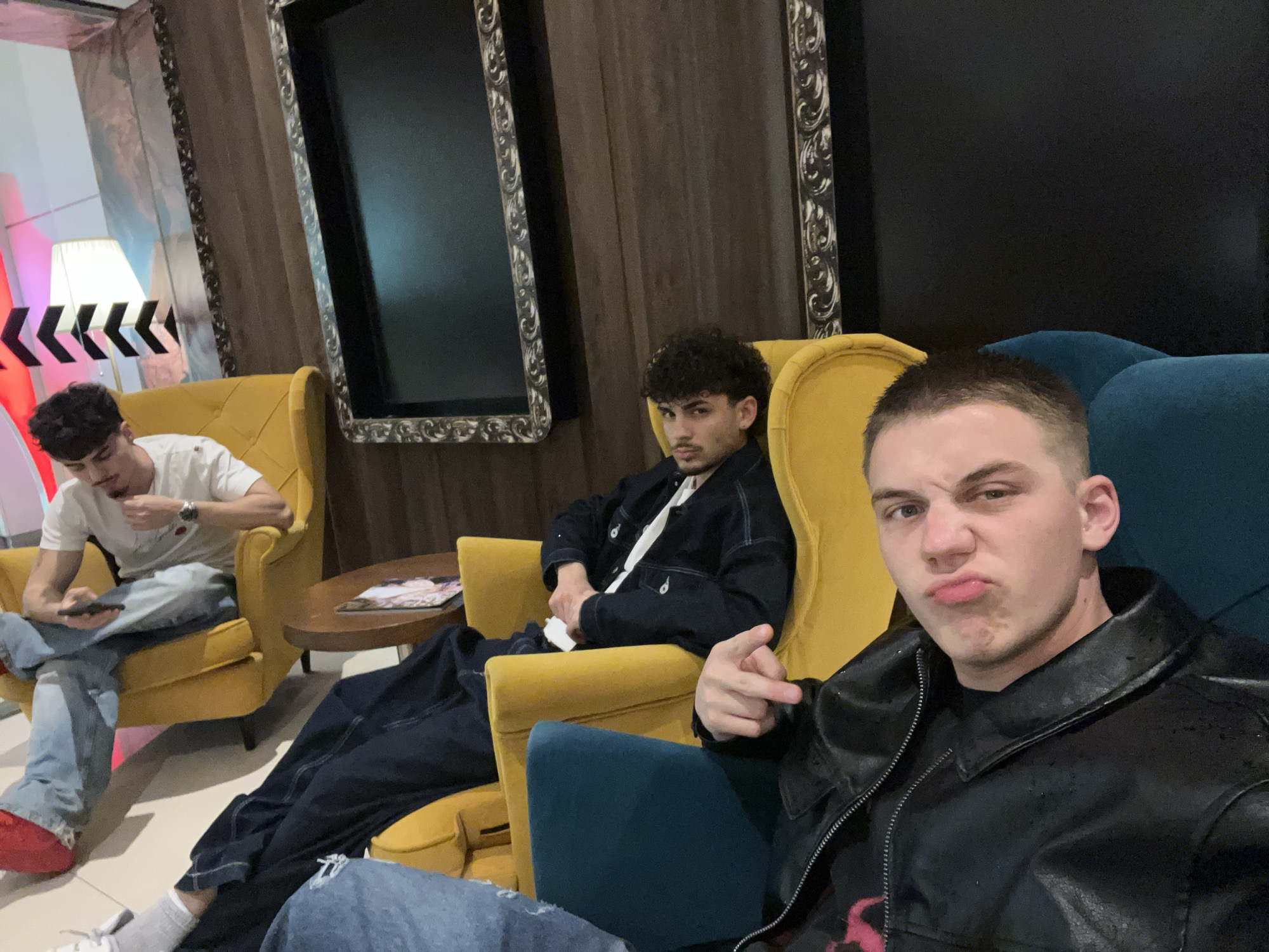 The image displays three young men relaxing in an indoor lounge or waiting area, possibly in Sofia, Bulgaria. The setting features two mustard-yellow armchairs and one blue armchair, suggesting a comfortable, modern environment with classic decorative touches.
In the foreground, positioned on the right, a light-skinned man with short, dark hair wears a black leather jacket over a black t-shirt. He looks directly at the camera with a playful, pouting expression and points a finger towards the viewer, indicating a selfie or direct interaction with the photographer. He is seated on the blue armchair.
To his left, in the mid-ground, a man with curly dark hair and a mustache sits in a yellow armchair. He is dressed in a dark denim jacket over a white t-shirt and looks intently at the camera with a more serious or neutral expression.
Further to the left, in the background, a third man with dark curly hair, wearing a white t-shirt and light blue jeans, is seated in another yellow armchair. He appears to be focused on his smartphone, which he holds in his hands, with one hand near his mouth. His feet, clad in red shoes, are visible.
Between the two yellow armchairs, a small round wooden coffee table holds an unidentifiable magazine or book. The background wall features two large, dark-surfaced mirrors or screens with ornate, silver-colored frames. To the far left, a floor lamp with a white shade and a distinctive black and white chevron pattern, reminiscent of a film clapperboard, is visible. The artificial lighting suggests the scene takes place during the evening or night. No clearly legible text is visible in the image.