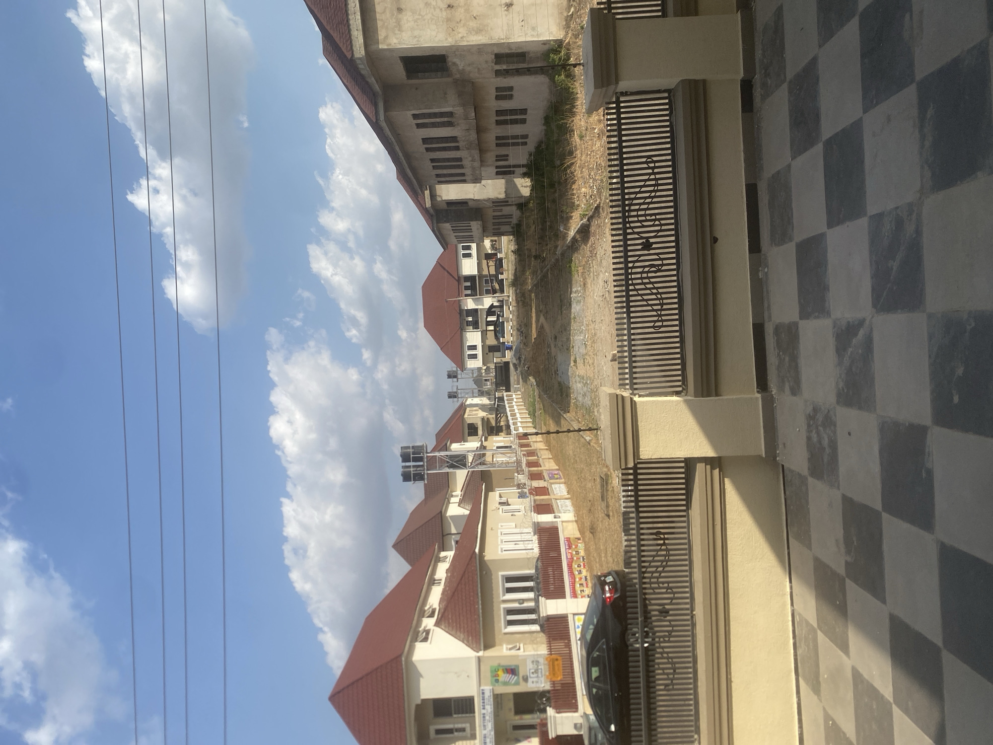 The image depicts a residential street in Iddo Tudunwada, Nigeria. Rows of two-story houses with red roofs line the street. The houses appear to be of similar construction and design. A black car is parked in front of one of the houses. Utility poles and wires are visible against a blue sky with white clouds. The foreground features a checkered sidewalk and a metal fence. The houses are mostly white/tan with brown roofing. There are water tanks at the tops of the houses and possibly solar panels.