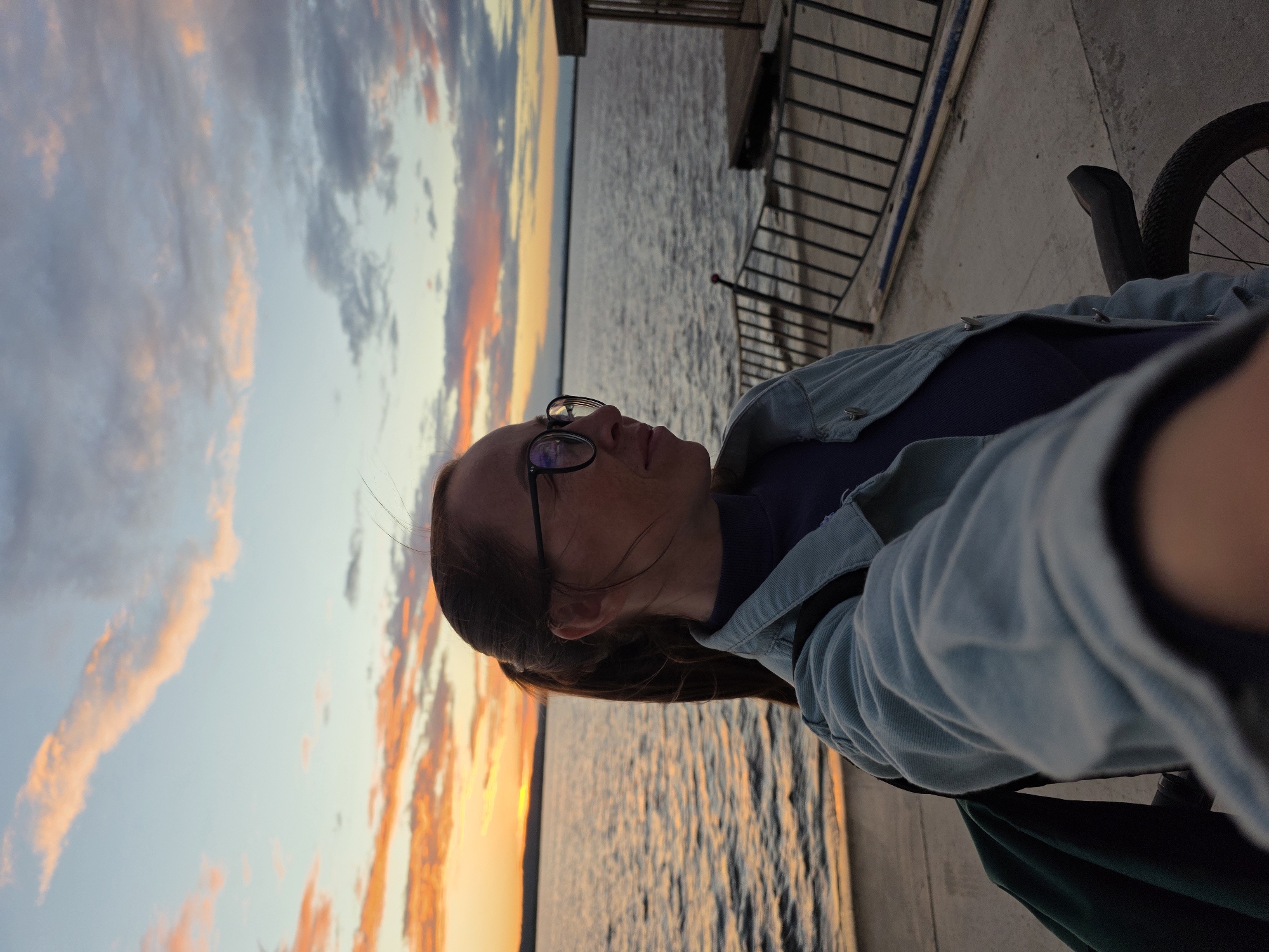 The media file shows a person on a paved surface near a body of water. The person is wearing glasses and a light blue denim jacket over a dark top. Their head is tilted upwards, looking towards a dramatic sunset. The sky is filled with clouds in shades of orange, pink, and blue. The water reflects the colors of the sky. To the right of the person, a bicycle is partially visible, with its wheel and frame in focus. Behind the person and to the right, there is a railing and a structure that appears to be a pier or boardwalk. The scene suggests a relaxed or contemplative moment during sunset, possibly after a bike ride. The lighting and cloud formations indicate it is late evening, during the golden hour. The location is Chaikovsky, Russia. There is no visible text in the image.