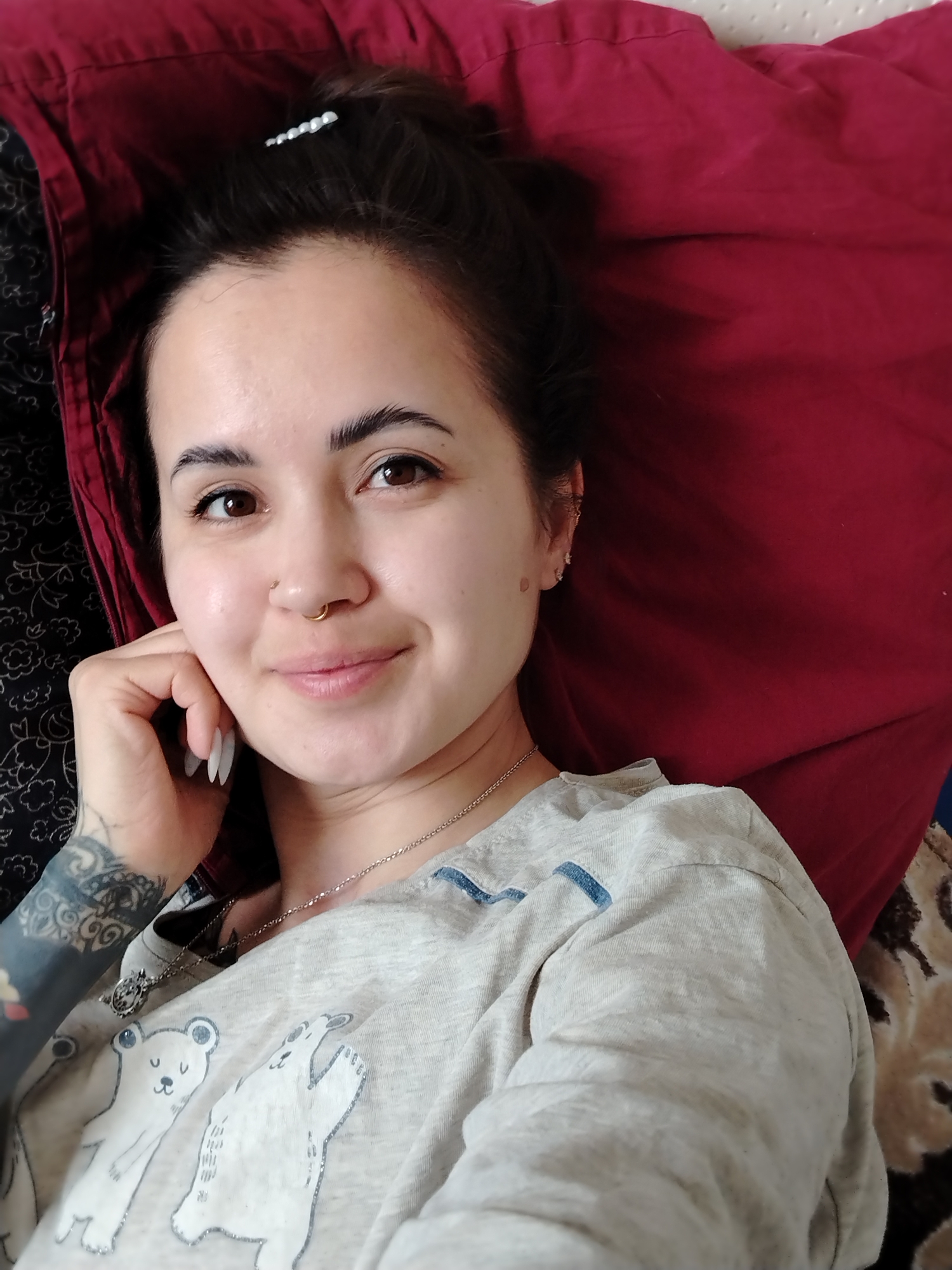 A close-up selfie shows a young woman lying down indoors, likely in Noviy, Russia, with her head resting on a dark red pillow or blanket. Her dark hair is pulled back, secured by a silver clip, framing her face. She has brown eyes and a subtle, pleasant smile. Visible on her face are a gold septum ring and a stud piercing on her right nostril. Her right hand, with long, white, pointed fingernails, is casually positioned near her cheek. She wears a light grey t-shirt featuring white illustrations of two bears and a silver necklace with a small pendant. Intricate dark tattoos are visible on her left arm. The scene conveys a relaxed and comfortable atmosphere.