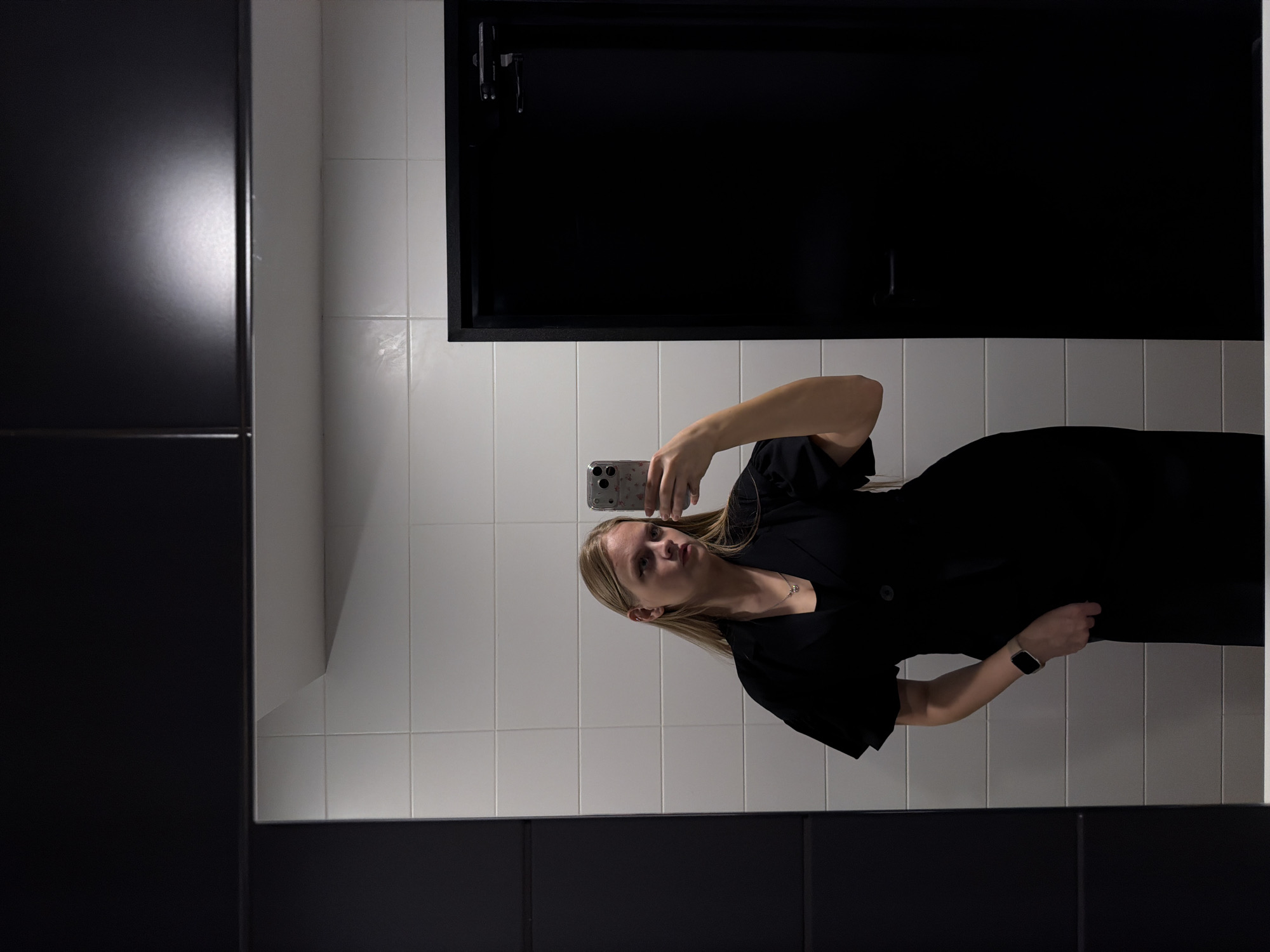 A young, blonde woman in a black dress stands in a bathroom taking a selfie in the mirror. The bathroom walls are covered in white tiles with black trim. Above her, there is a black cabinet. A possible location in Izhevsk, Russia is indeterminate from the given details.