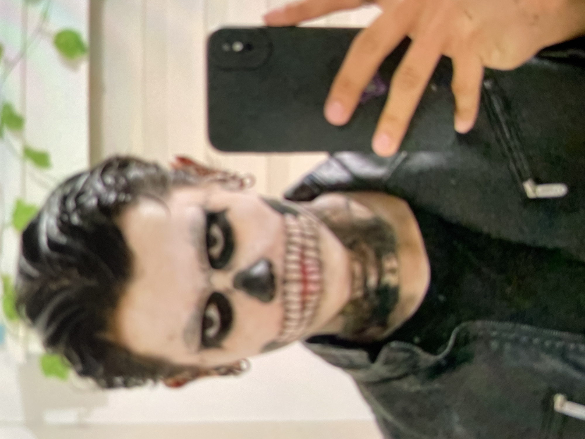 A person with skull makeup is taking a selfie. The person has pale skin, black eye sockets and a nose painted black. There are also teeth painted onto the lower half of the face in red and white. The person is wearing a black leather jacket and holding a black smartphone. The background appears to be a wall with some plants. The location is likely indoors. There is no information to verify the location is in Harmanli, Bulgaria.