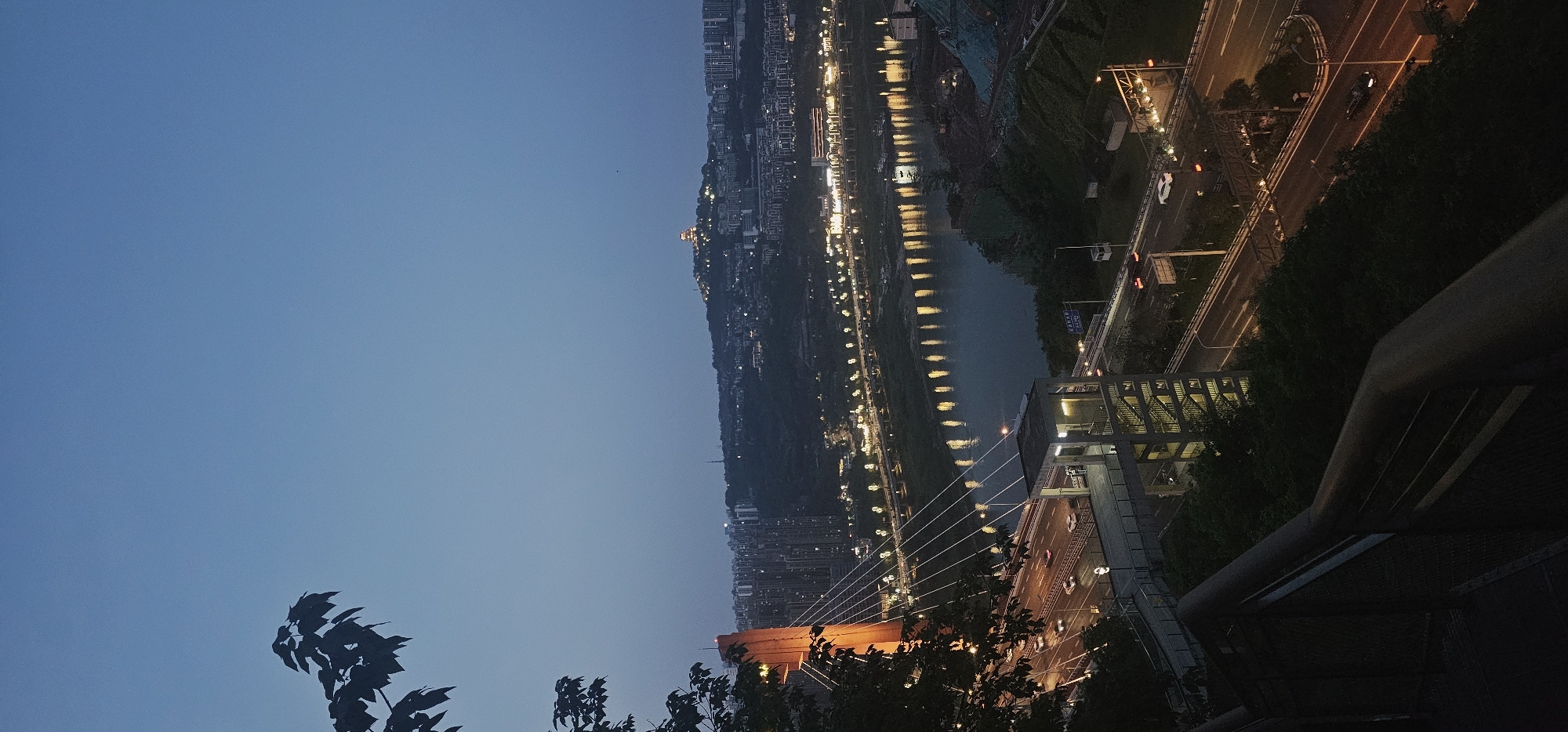 View of Yuzhong District, China, at dusk. The sky is a gradient of blue, transitioning to a darker hue closer to the horizon. A body of water reflects the city lights, with a line of buildings and illuminated streets running parallel to the water. A bridge with orange support structures is visible in the foreground. Vegetation is visible along the roadside and in the foreground. Faint traffic is seen on the roads. A portion of a railing is also visible in the foreground.