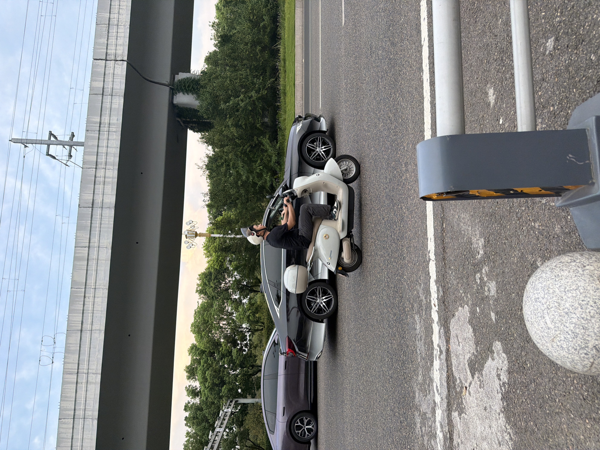 In Jiangjin, China, a man wearing a helmet rides a light-colored scooter down a paved road. He is positioned in the center of the frame, in front of a dark-colored sedan and a purple sedan, both of which are on the road. Overhead, a concrete overpass looms, with power lines visible against a hazy sky. The road surface is asphalt, marked with white lines. To the right of the scooter, a grey and yellow barrier is partially visible. Trees line the background beyond the road.
