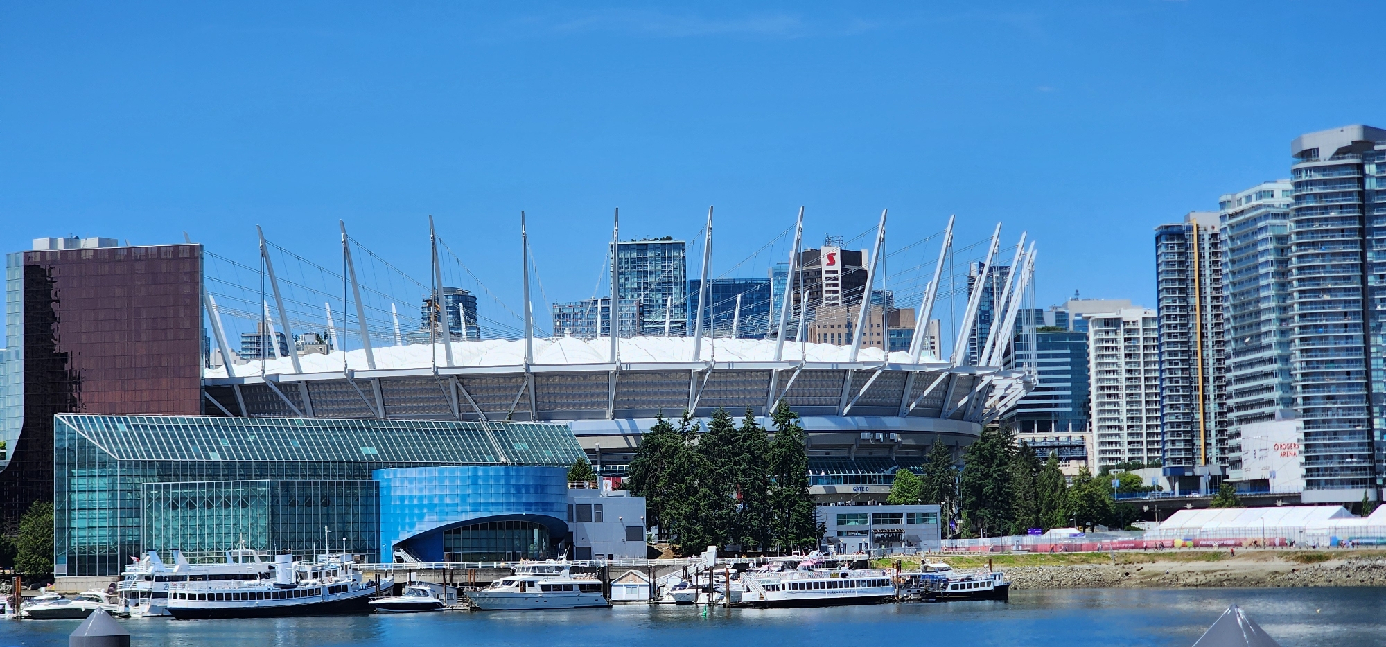 This is a medium-angle, eye-level, exterior shot of a modern stadium and surrounding cityscape on a clear, sunny day. The stadium, with its distinctive white cable-supported roof, dominates the center of the frame. In front of the stadium, a contemporary building with a large glass facade and a curved blue section is visible. Several boats are docked in the foreground on the water. Behind the stadium, a dense cluster of high-rise buildings, primarily glass and concrete, forms the urban backdrop. The sky is a vibrant, cloudless blue. The scene suggests an urban environment with a focus on modern architecture and a waterfront setting.
