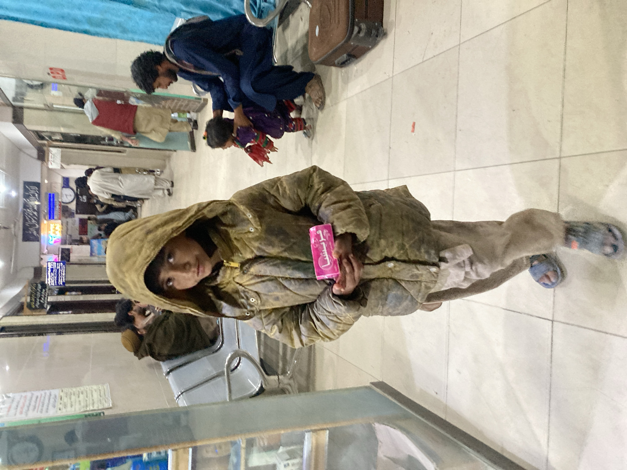 The image shows a young person standing in what appears to be a public waiting area. The individual is wearing a hooded jacket and holding a pink package with writing in Arabic. The setting includes tiled flooring and seating areas with other individuals present. In the background, there are signs with Arabic script, indicating a location in Quetta City Tehsil, Pakistan. Other people are seen with luggage suggesting an area where people may be arriving or departing.