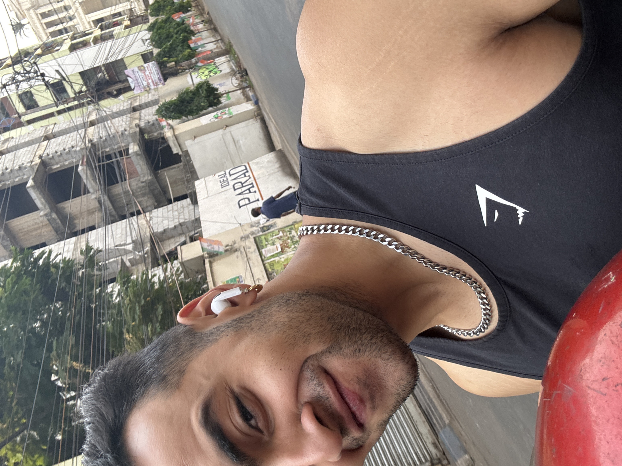 A close-up, upward-angled selfie features a light-skinned man with short, dark hair and stubble, wearing a black tank top with a white logo. He has a silver chain around his neck and a white wireless earbud in his left ear.
The background shows an urban street scene in Kolkata, India, with buildings under construction and a street with visible signage. A man in a dark blue shirt walks in the background. Wires are strung across the buildings.
