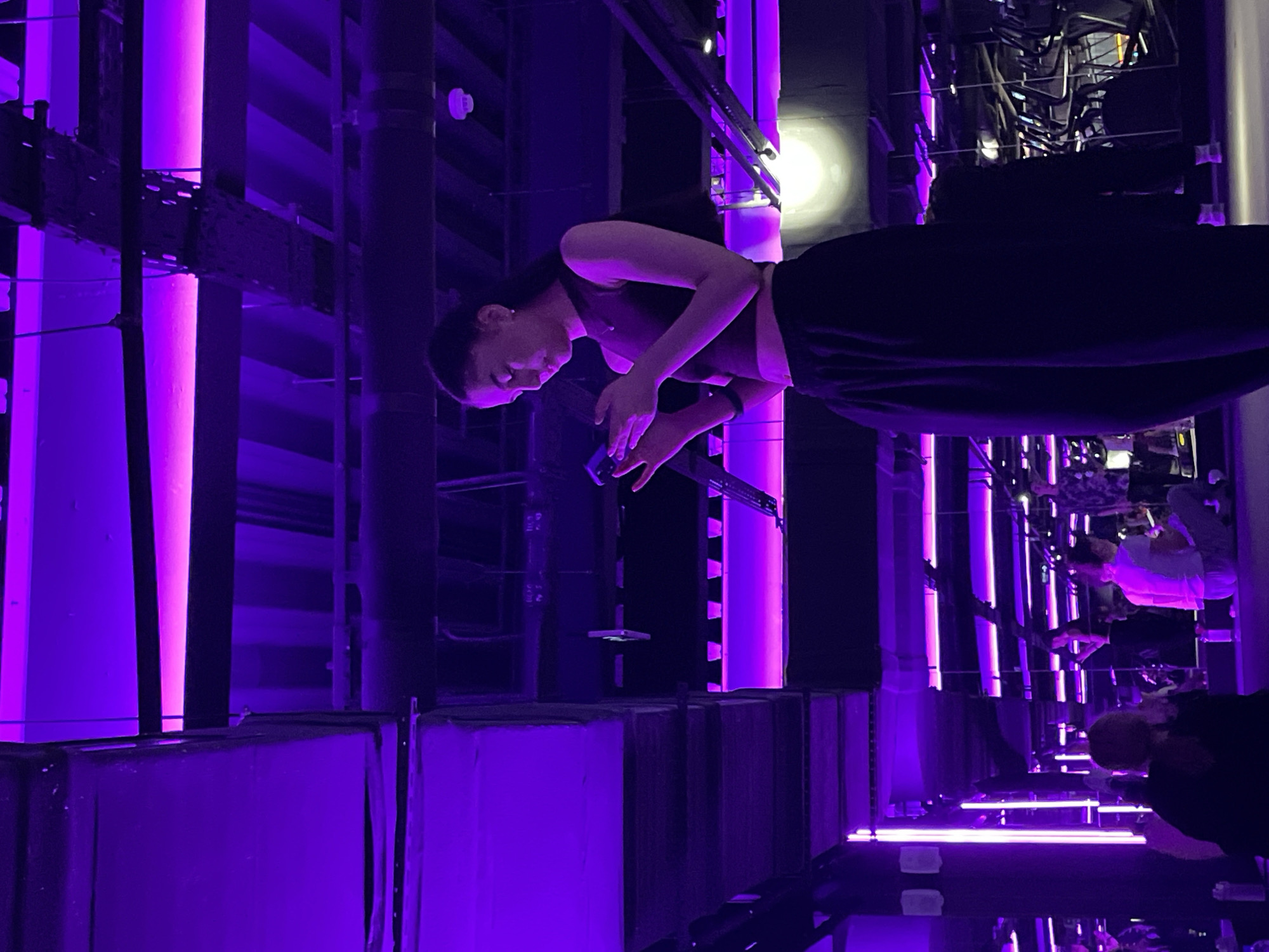 A woman in a dark sleeveless top and a dark, full skirt is looking down at a mobile phone held in her hands. She is standing in a room with reflective surfaces and linear lighting in shades of purple and white. The room is lined with black ducts or boxes. There are reflections of other people in the background. The lighting casts a strong, vibrant purple hue throughout the scene. The location is the Museum of Illusions in Saint Petersburg, Russia.