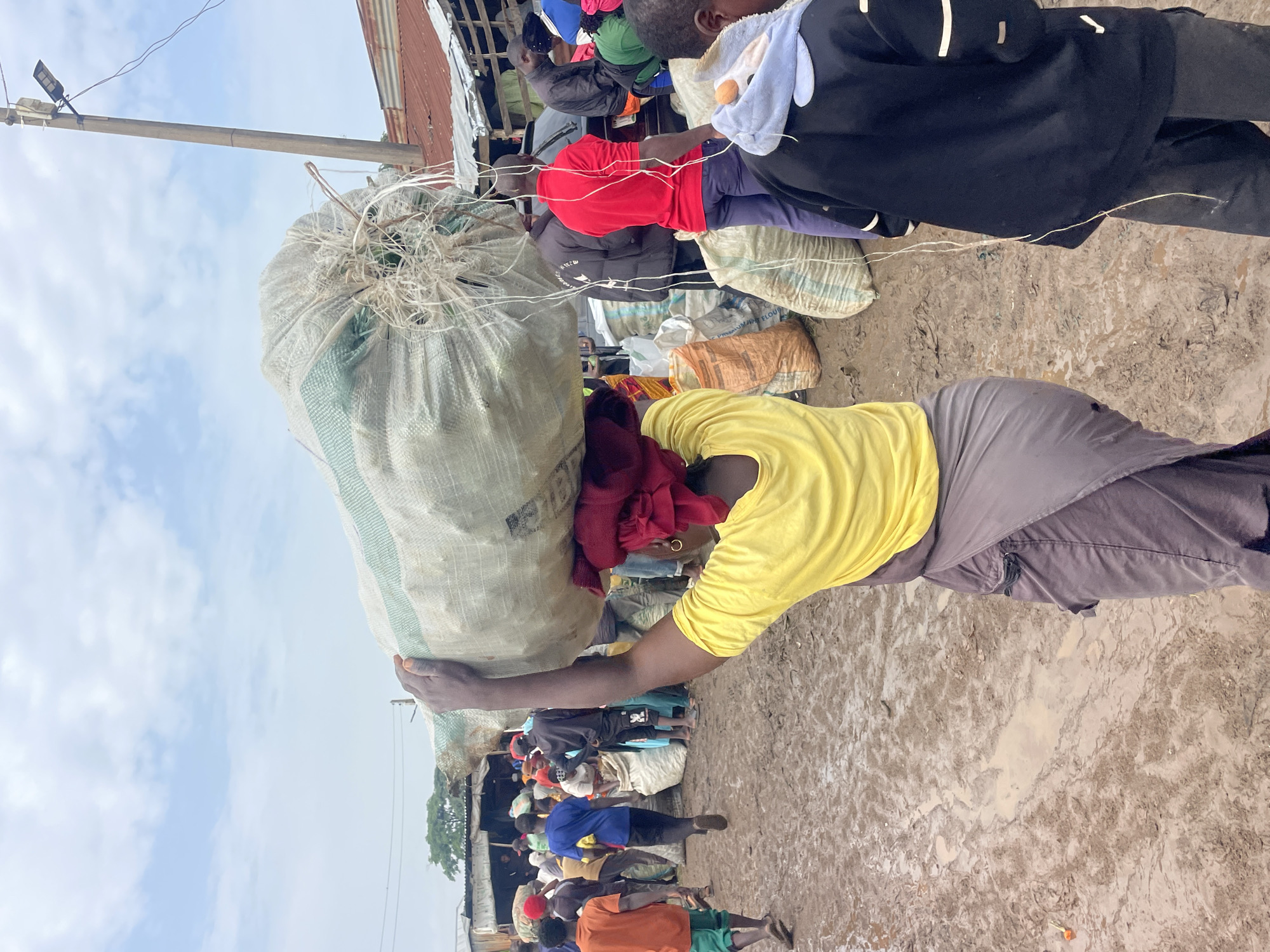 In Gura Top, Nigeria, a woman carries a large, light-colored burlap sack of goods on her head. She wears a yellow t-shirt and a purple wrap skirt, with a red cloth tied around her head. The scene is a bustling outdoor market with muddy ground, suggesting recent rainfall. Several other people are visible, some carrying sacks and others milling about. Stalls with corrugated metal roofs are present in the background, along with a utility pole and overhead wires. The sky is cloudy.