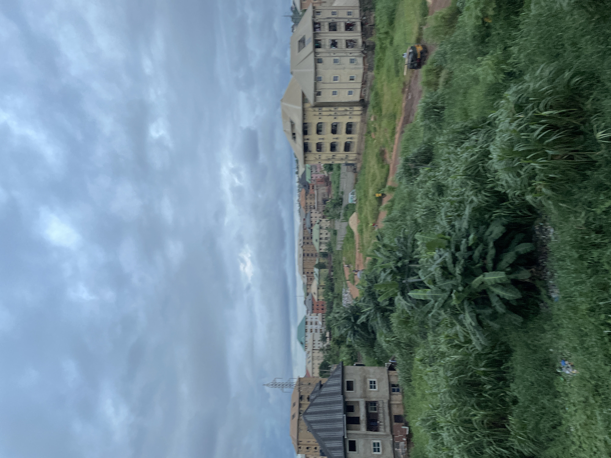 This is a wide shot of Nkpor, Nigeria, showing a residential area under a cloudy sky. In the foreground, lush green vegetation, including banana trees and tall grass, dominates the landscape. A two-story building with a gray, corrugated roof and white-framed windows is visible on the left.
Further back, a dirt road winds through the scene. Several multi-story buildings, appearing to be apartment complexes, are scattered across the middle ground and background. One building stands out with a distinctive green roof. A power pylon is visible in the distance. A yellow and black three-wheeled vehicle, possibly a tuk-tuk or a small utility vehicle, is parked on the dirt road. A few individuals are visible as small figures on the road and near the buildings, suggesting ongoing activity in the area. The overall impression is of a developing urban environment with a mix of residential structures and natural vegetation.