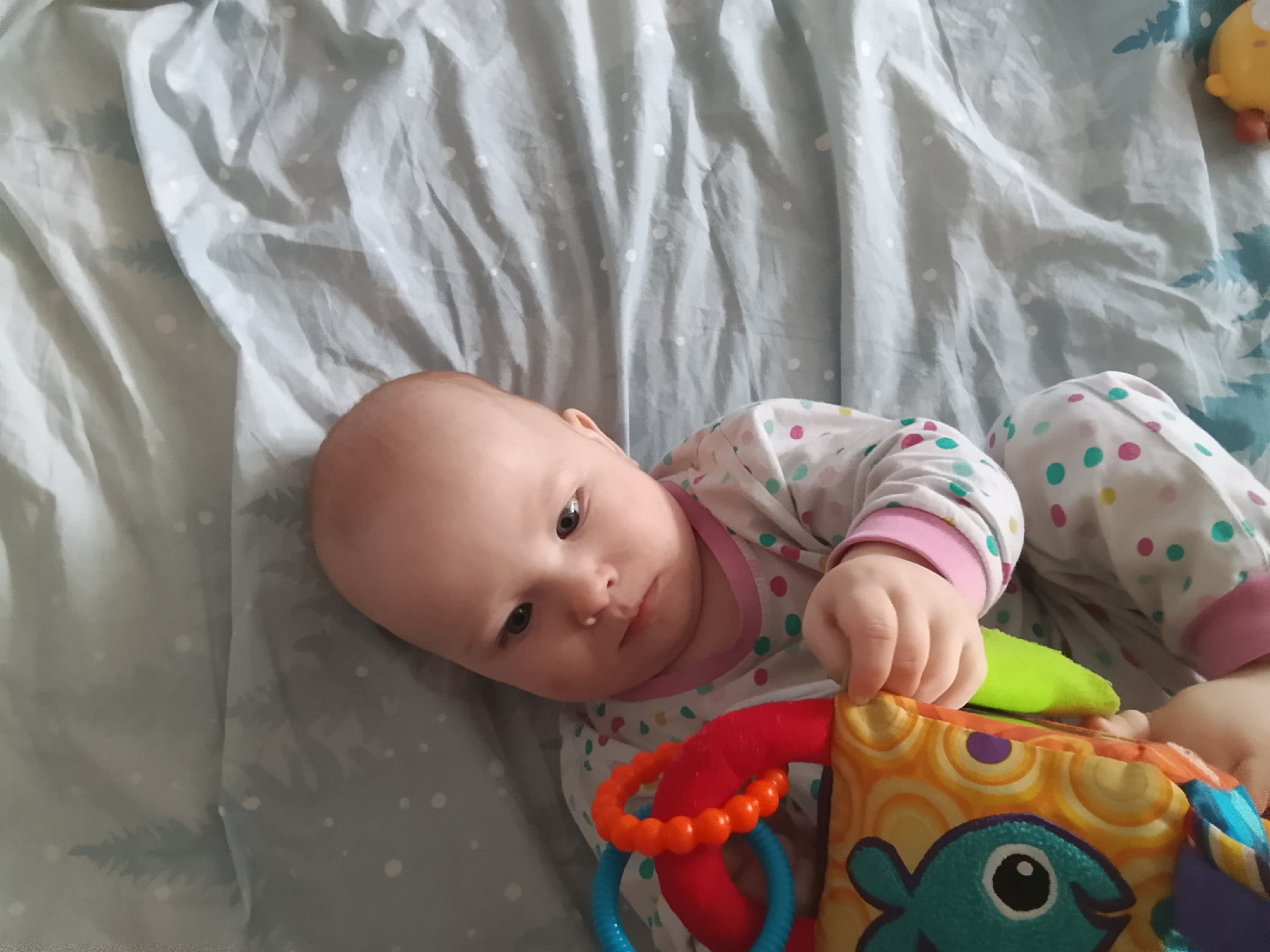 A close-up, high-angle shot shows a baby lying on a gray, wrinkled sheet with a subtle white polka dot pattern. The baby is wearing a white pajama set adorned with colorful polka dots in pink, green, and turquoise, with pink trim on the cuffs. The baby's head is turned to the left, with their gaze directed towards the camera. The baby's left hand is resting on a colorful soft book, which features a design with an orange border, concentric circles, and a turquoise fish with a black eye. Attached to the book are red and blue plastic rings. A bright green fabric object, possibly a part of the toy, is visible to the left of the book. In the upper right corner, a yellow toy with orange accents is partially visible. The overall scene suggests a domestic setting, likely a nursery or bedroom. There is no information available to determine the location in the city of Minsk, Belarus.