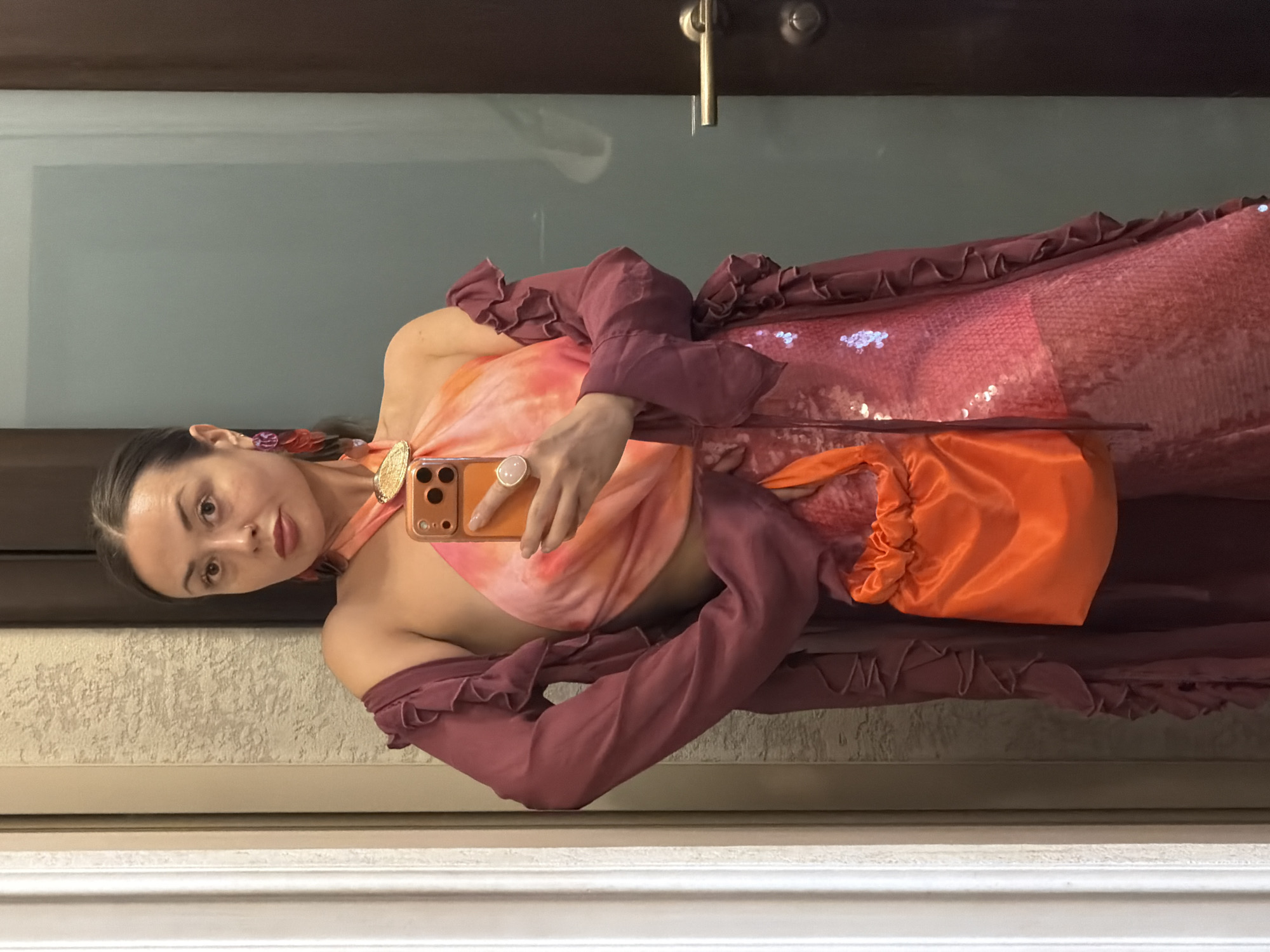 A woman stands in front of a mirror, taking a selfie. She is wearing a vibrant outfit consisting of a pink sequined dress and a sheer, ruffled burgundy kimono. She accessorizes with large, dangling purple earrings, a gold pendant necklace, and a bright orange drawstring pouch bag. A pink ring is visible on her right hand. The background appears to be an interior space, possibly a home or hotel room, with textured walls and a large mirror. A door with a silver handle is visible to the right. There is no indication that this image was taken in Krasnoyarsk, Russia.