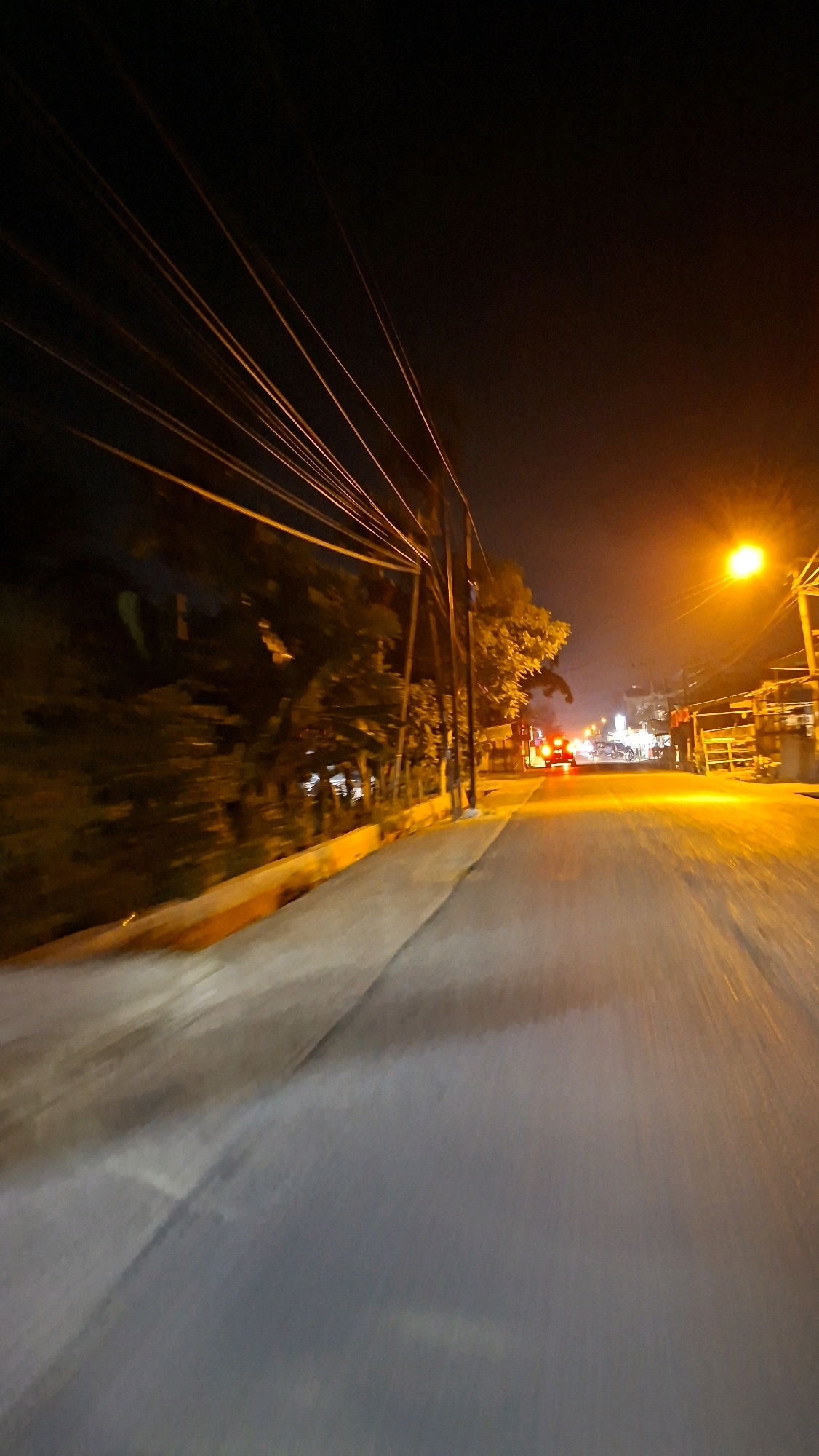 The image depicts a nighttime scene on a road in Lhokseumawe, Indonesia. The perspective is from a moving vehicle, suggesting a travel or transportation context. The road is paved, with the right side illuminated by a bright street lamp casting an orange glow. On the left, trees and vegetation line the road, with some fencing visible. Overhead, a dense network of electrical wires stretches across the frame, contributing to the urban feel. In the distance, the red taillights of a vehicle are visible, indicating traffic. The overall ambiance is one of a typical street scene at night in an Indonesian city, with the lighting and infrastructure suggesting a developed area.