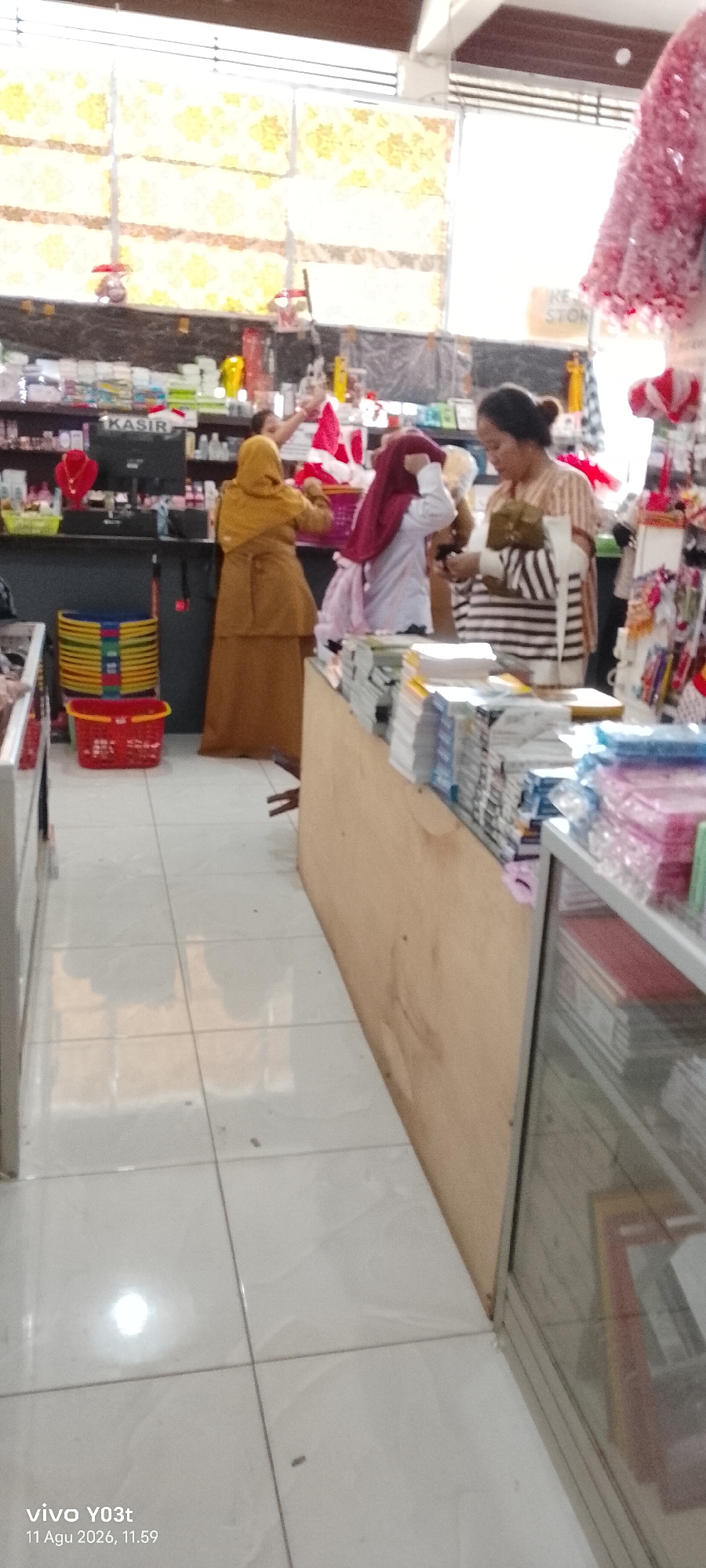 The image shows the interior of a store, likely a small shop or market, located in Atula, Indonesia. The store appears to sell a variety of goods, including stationery, toiletries, and possibly clothing accessories.
In the foreground, a floor made of light-colored square tiles is visible. The tiles show reflections, indicating lighting from above.
In the mid-ground, there are several people. On the left, a woman in a yellow hijab and a long, earth-toned dress is standing behind a counter. She appears to be attending to something on the counter. To her right, another person, mostly obscured, is also behind the counter, interacting with items. Further to the right, a woman wearing a red hijab and a white top is seen interacting with a basket of items. In the far right of the mid-ground, a woman with dark hair pulled back is wearing a striped shirt and appears to be looking at items in her hands.
Various objects are visible in the store. Behind the counter on the left, shelves are stocked with numerous products, including bottles and boxes. A sign that says "KASIR" (cashier in Indonesian) is visible on the counter. Several colorful plastic baskets are stacked on the floor to the left of the counter, along with a red plastic basket. In the background, more shelves with products are present, and a decorative pink item, possibly clothing or fabric, hangs from the ceiling on the far right.
The environment suggests a retail space with merchandise displayed on shelves and counters. The lighting appears to be artificial, possibly from fluorescent lights, and there's a bright reflection on the floor. The overall impression is of a busy, functional retail environment.