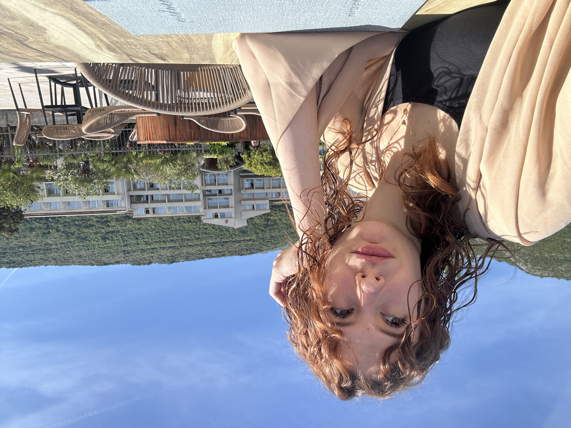 The image is a selfie taken outdoors in Petrovac centar, Montenegro. The subject is a woman with red curly hair, wearing a black top and a light beige cover-up. She is positioned with her head tilted, looking at the camera. Behind her, the sky is visible, transitioning to a green hillside and a building with multiple stories. In the upper portion of the image, there is a table and chairs, suggesting an outdoor dining area.