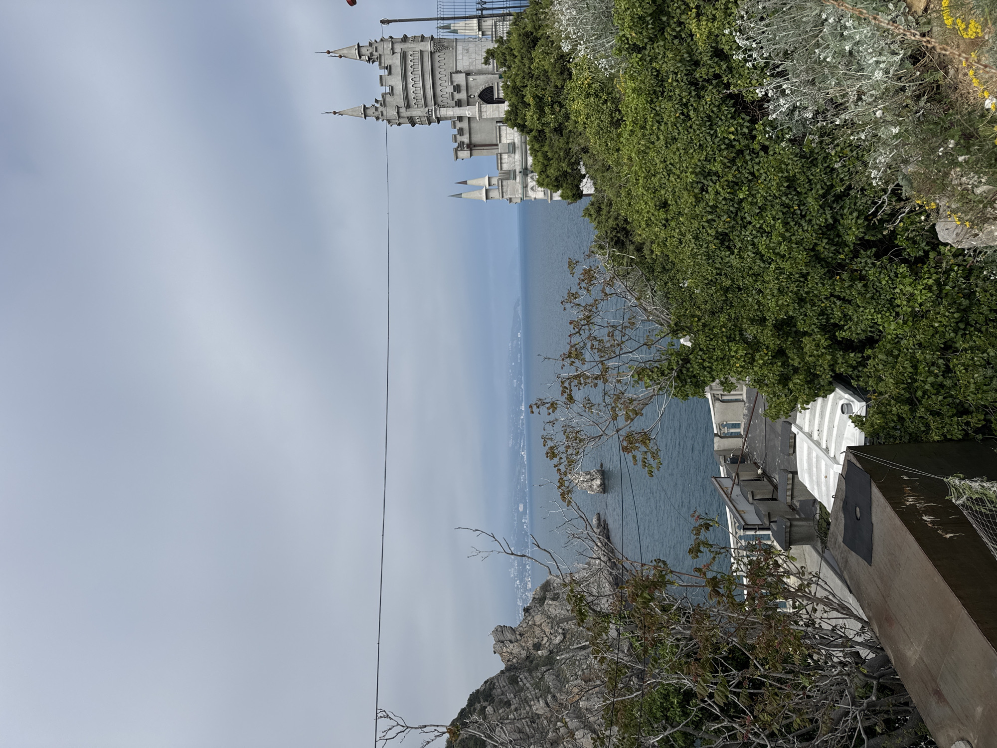 The image shows a sideways view of Swallow's Nest castle in Гаспринский поселковый совет, Ukraine, situated on a cliff overlooking the Black Sea. The castle, a prominent white structure with turrets, is partially obscured by green vegetation. Below the castle, the sea extends towards the horizon. In the foreground, there are rocky cliffs and foliage. A power line cuts across the view. Buildings and structures are visible on the cliffside.