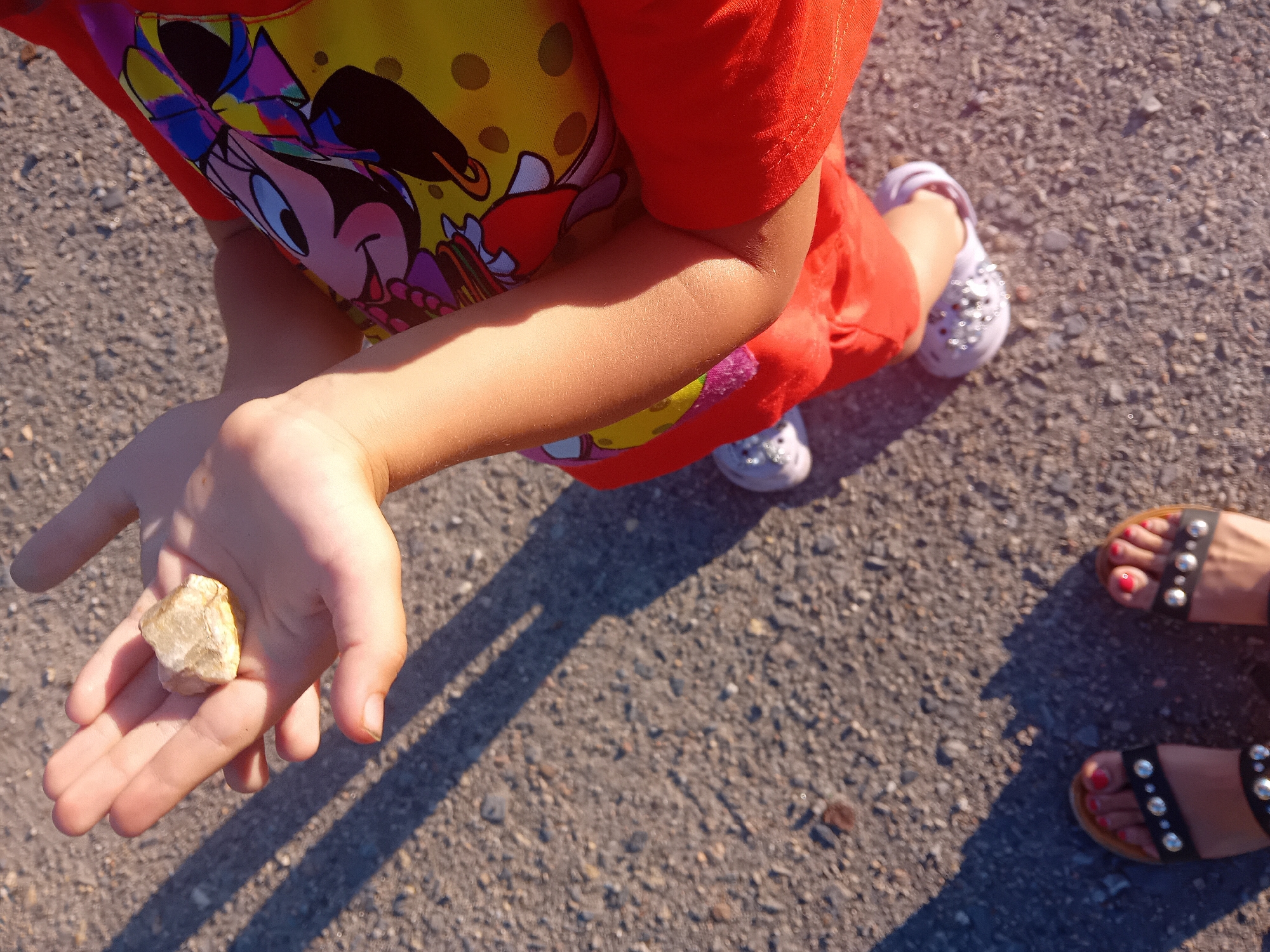 A child's hand holds a light-colored rock. The child wears a yellow shirt with a colorful print of Minnie Mouse and orange pants. Their feet are shod in white sandals. In the foreground, a pair of feet wearing black sandals with silver studs are visible. The scene is set on a paved surface, possibly a street or sidewalk in Chuhuiv, Ukraine. Shadows from the people suggest it is daytime.