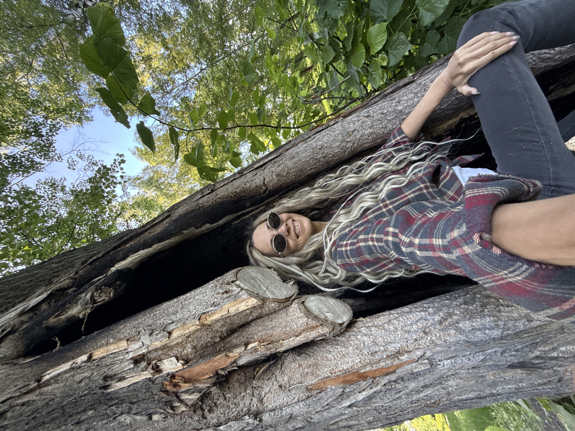 The image features a young woman with long, wavy blonde hair, lying inside a large, hollowed-out tree trunk in an outdoor, treed environment. She is wearing round sunglasses, a red, blue, white, and brown plaid flannel shirt, and dark pants. The woman is smiling broadly towards the camera, conveying a happy and relaxed mood. Her left arm is bent, resting on her left leg, while her right arm extends upward along the trunk.
The tree trunk she is resting in is substantial, with rough, textured bark on its exterior and a dark, weathered, and somewhat charred interior, suggesting it might be an old, fallen log. The perspective of the photograph is from a low angle, looking up past the woman and the log towards a clear blue sky, which is visible through a dense canopy of vibrant green leaves and tree branches. The scene is brightly lit by natural daylight, indicating it is daytime. The setting suggests a natural area like a forest or park, located in Rusanovo, Russia. There is no visible text in the image.