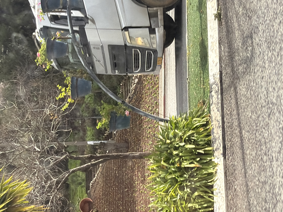 The image depicts a partial view of a light-colored Volvo truck on an asphalt road in Safed, Israel. A prominent dark, curved metal arm extends from the truck's front right side, from which three dark blue plant pots are suspended at various heights. The pots contain green foliage, with pink flowers visible in two of them. The truck's front grille, headlights, and part of its front wheel are visible, along with a yellow license plate. Adjacent to the road is a concrete curb and a grassy strip, followed by a dense bush of long, green leaves. In the background, a hillside rises, featuring a bare-branched tree, terraced stone steps, and an empty terracotta pot. The hillside is sparsely covered with green undergrowth and fallen leaves. The scene is illuminated by natural daylight, casting shadows on the road. No people are visible.