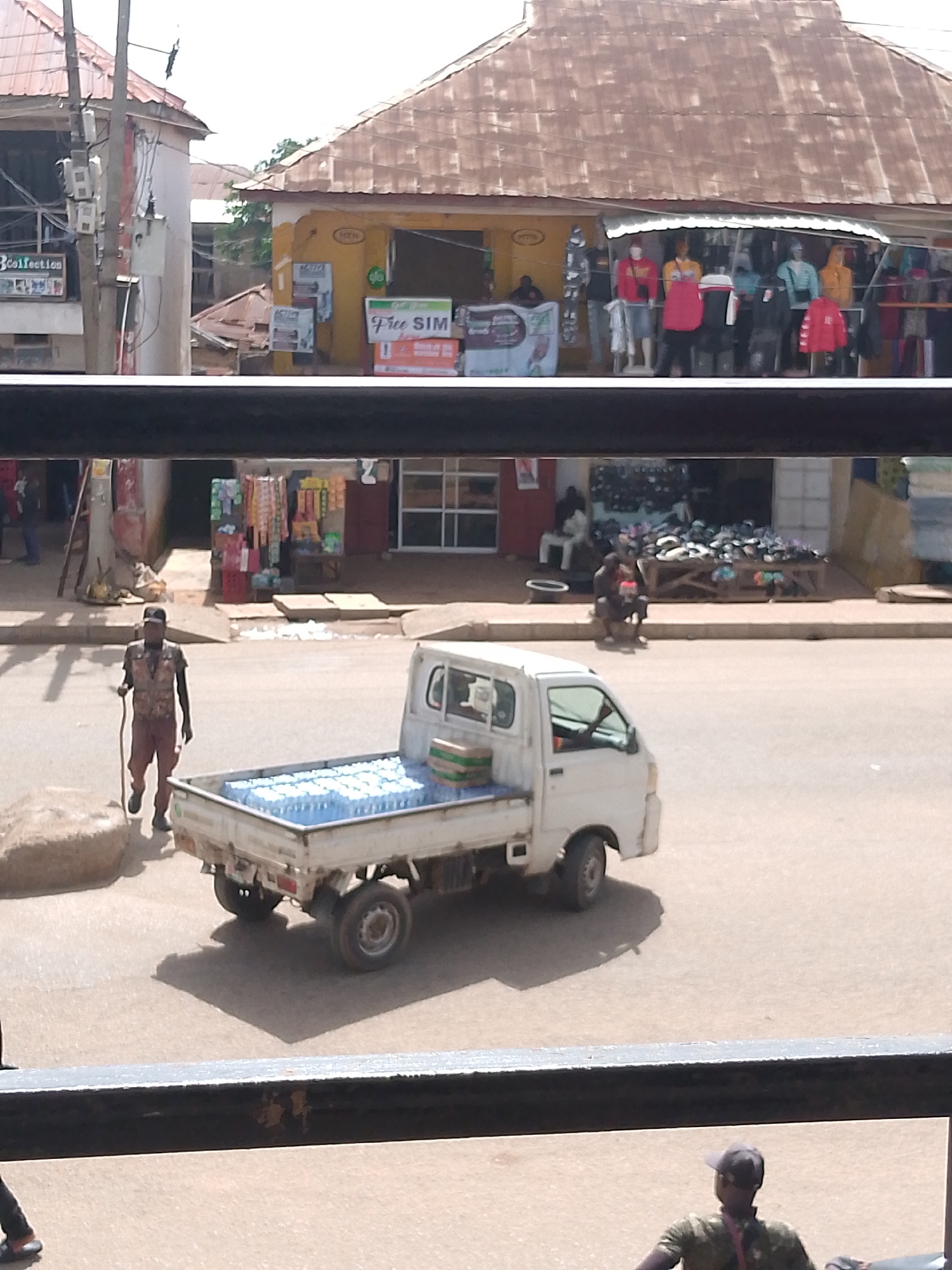The image captures a street scene in Bukuru, Nigeria, with a focus on a small white pickup truck loaded with bottled water. The truck is parked on a paved road, and its bed is filled with numerous clear plastic bottles of water, some stacked in green crates. To the left of the truck, a man wearing a dark vest over a long-sleeved shirt and dark pants, carrying a walking stick, walks away from the truck. He has a dark cap on his head. In the background, there is a row of shops. One shop, painted yellow, has displays of clothing on mannequins and a sign advertising "Free SIM". Other shops appear to be selling various goods, including what looks like food items and shoes. The architecture suggests a bustling market area with corrugated metal roofs on some buildings. The overall lighting indicates it is daytime, with shadows cast on the road.