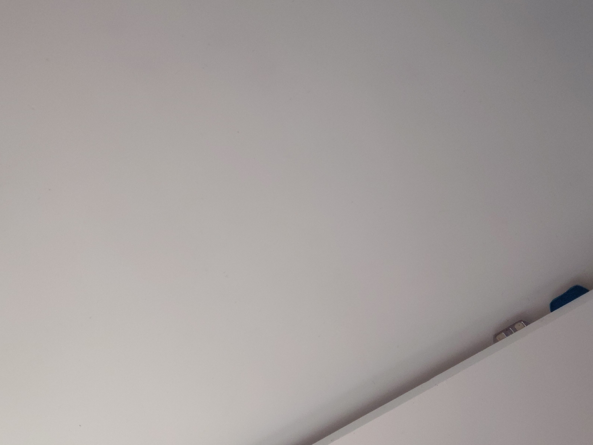 The image primarily depicts a plain, light grey or off-white surface, likely an interior wall or ceiling, occupying most of the frame. This surface is smooth with minor textural variations and a few small, dark specks. A sharp diagonal line delineates the lower right portion of the image, indicating the edge of another white surface, possibly a shelf or cabinet.
Along this diagonal edge, in the bottom right corner, several small, rectangular, metallic objects are visible. These appear to be silver-colored magnets or clips, suggesting they are affixed to the upper light-colored surface or the edge of the shelf. Further to the right, mostly obscured by the shelf edge, is a small, dark blue object, which could be a clip, a piece of plastic, or another small item.
The scene is static, with no people or discernible activity taking place. The lighting is even and diffused, consistent with an indoor environment. There are no indications of time of day, weather conditions, or specific features that would directly identify the location within Santa Ana, Venezuela. No visible text is present in the image.