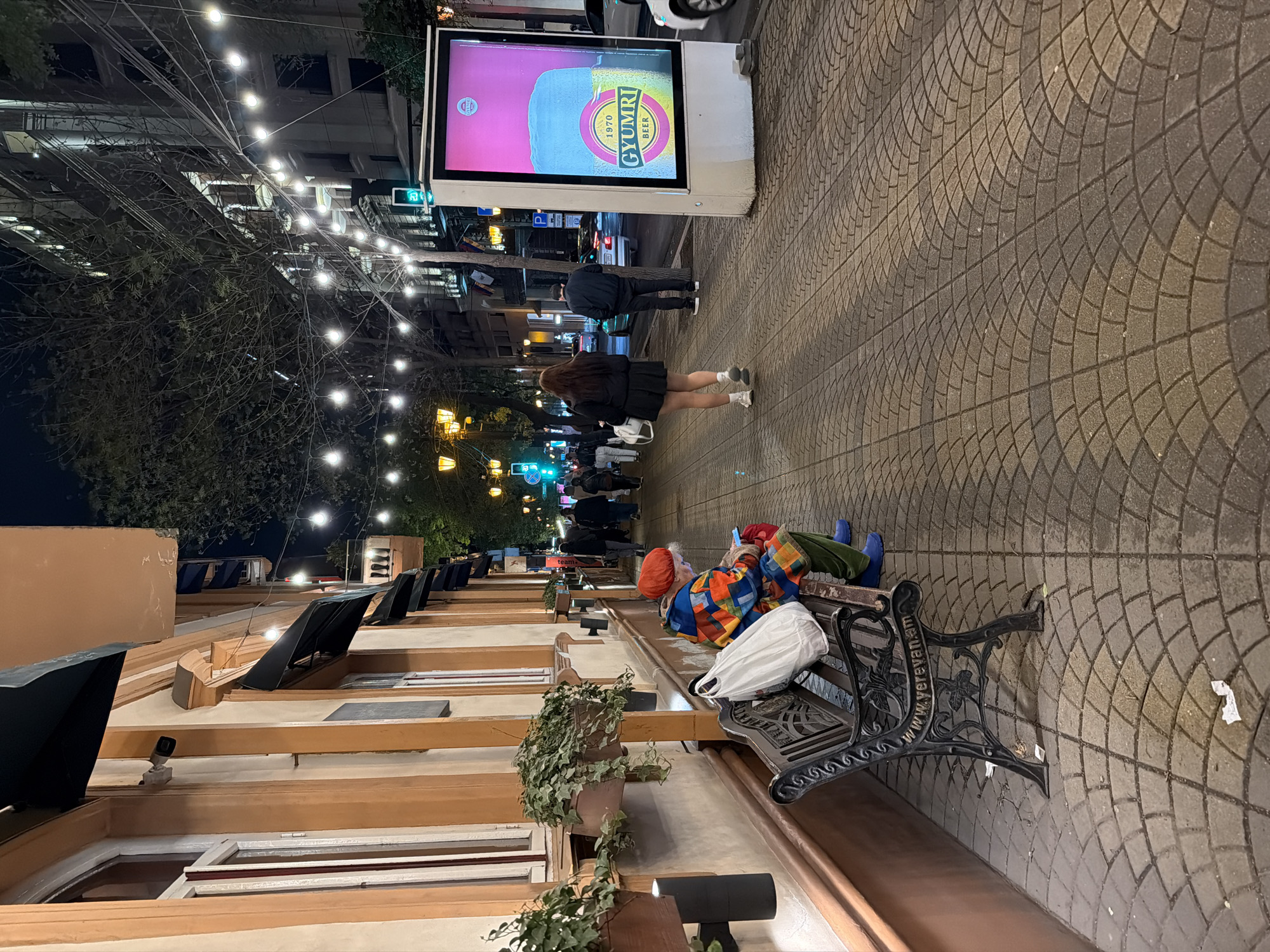 The image captures a vibrant nighttime scene on a pedestrian street in Yerevan, Armenia. The street is paved with intricately patterned cobblestones, and string lights are strung overhead, casting a warm glow and illuminating the path. Buildings with awnings and potted plants line one side of the street, suggesting commercial establishments.
In the foreground, a person is seated on a decorative black metal bench, distinctive with the inscription "www.yerevan.am". This individual wears colorful, patchwork-style clothing, an orange hat, and blue boots, appearing to be engrossed in a mobile device. A white bag rests beside them on the bench.
Further down the street, several other people are visible, including two individuals walking away from the camera. One is dressed in a dark jacket and pants, while the other (a woman) wears a black skirt and jacket. A prominent, illuminated vertical billboard stands out in the mid-ground, displaying an advertisement for "GYUMRI BEER" with the text "1970". A green traffic light is also visible nearby. The background shows more street activity and distant lights, indicating a bustling urban environment after dark.