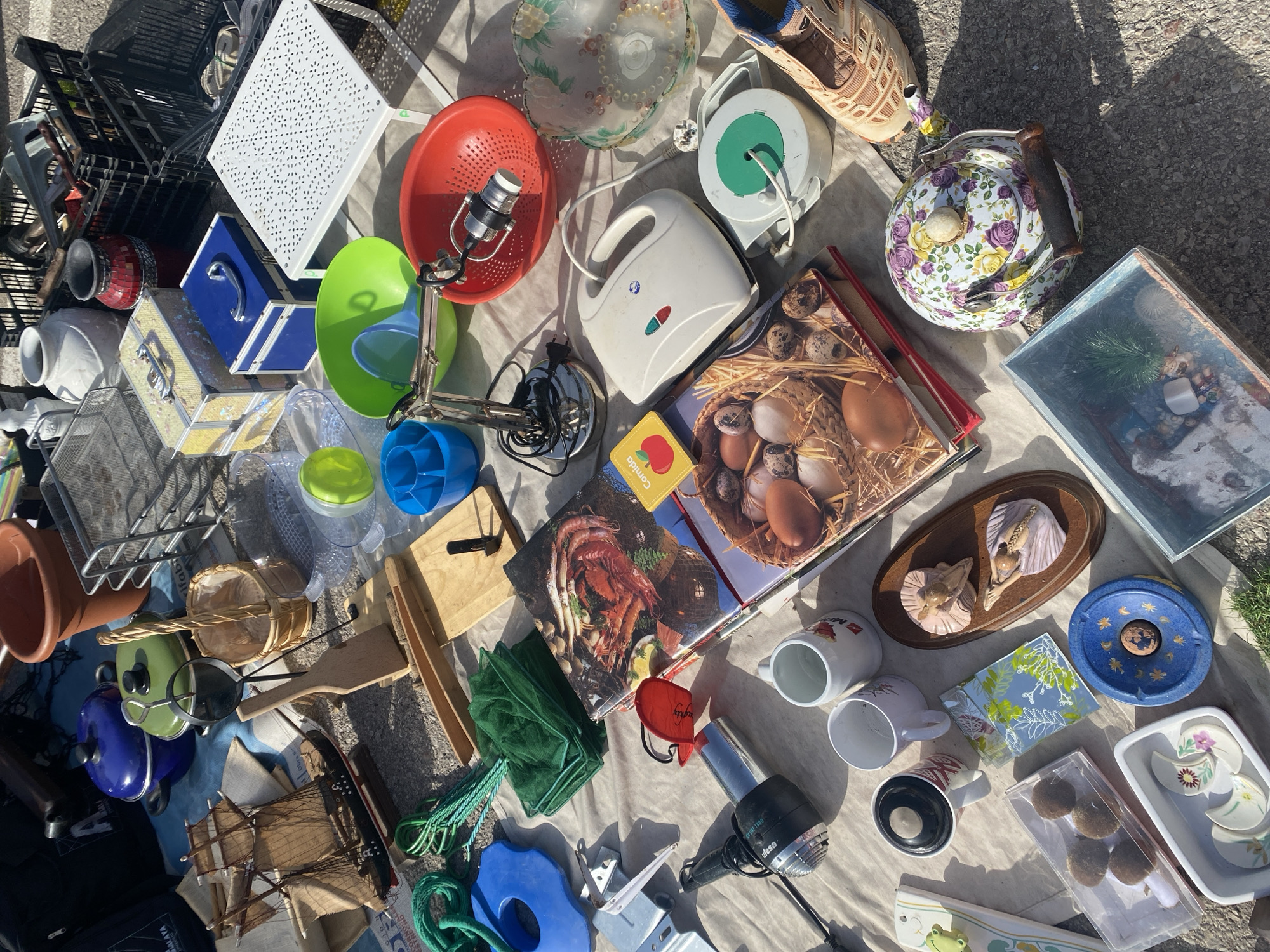 The image depicts an outdoor market stall, likely a flea market, with a variety of items laid out on a beige cloth and the ground. The scene is set on a sunny day, with harsh shadows indicating strong sunlight.
Prominently displayed are household items, decorative objects, and some tools. Among the notable items are a miniature sailing ship, several baskets (one of which is woven and appears to hold small items), ceramic pots, a red colander, a white sandwich maker, and a white extension cord reel. There are also various books, including one with an image of seafood and another with a close-up of eggs in a basket. Decorative pieces include a floral patterned teapot, a blue ashtray with celestial motifs, and small ceramic figurines. Kitchenware such as mugs and a glass bowl are also visible. Several green nets and a blue plastic object are on the left side of the image. Towards the top, there are blue and green pots, and metal shelving units.
The location is identified as Manilva, Spain. The ground surface appears to be a mix of gravel and asphalt, typical of outdoor public spaces or market areas. There are no people visible in the image. The arrangement suggests a retail or resale environment where items are presented for sale.