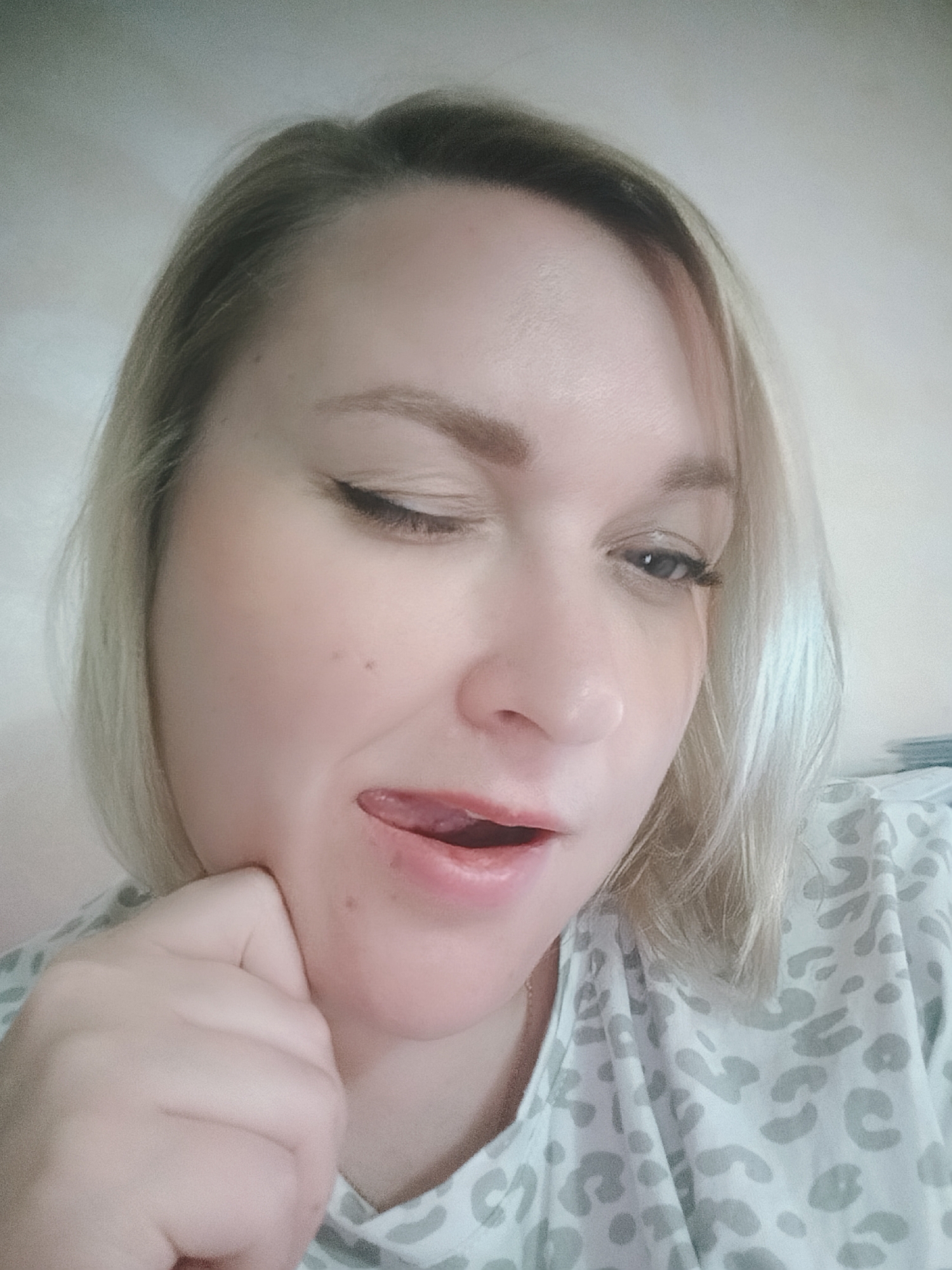 The image is a close-up self-portrait of a woman with blonde hair. She is looking down and to the side, with her mouth slightly open and tongue visible. One hand is raised to her chin. She has fair skin with a few visible moles. She is wearing a white shirt with a light gray animal print. The background is a plain, light-colored wall. The location is Unknown, Unknown.