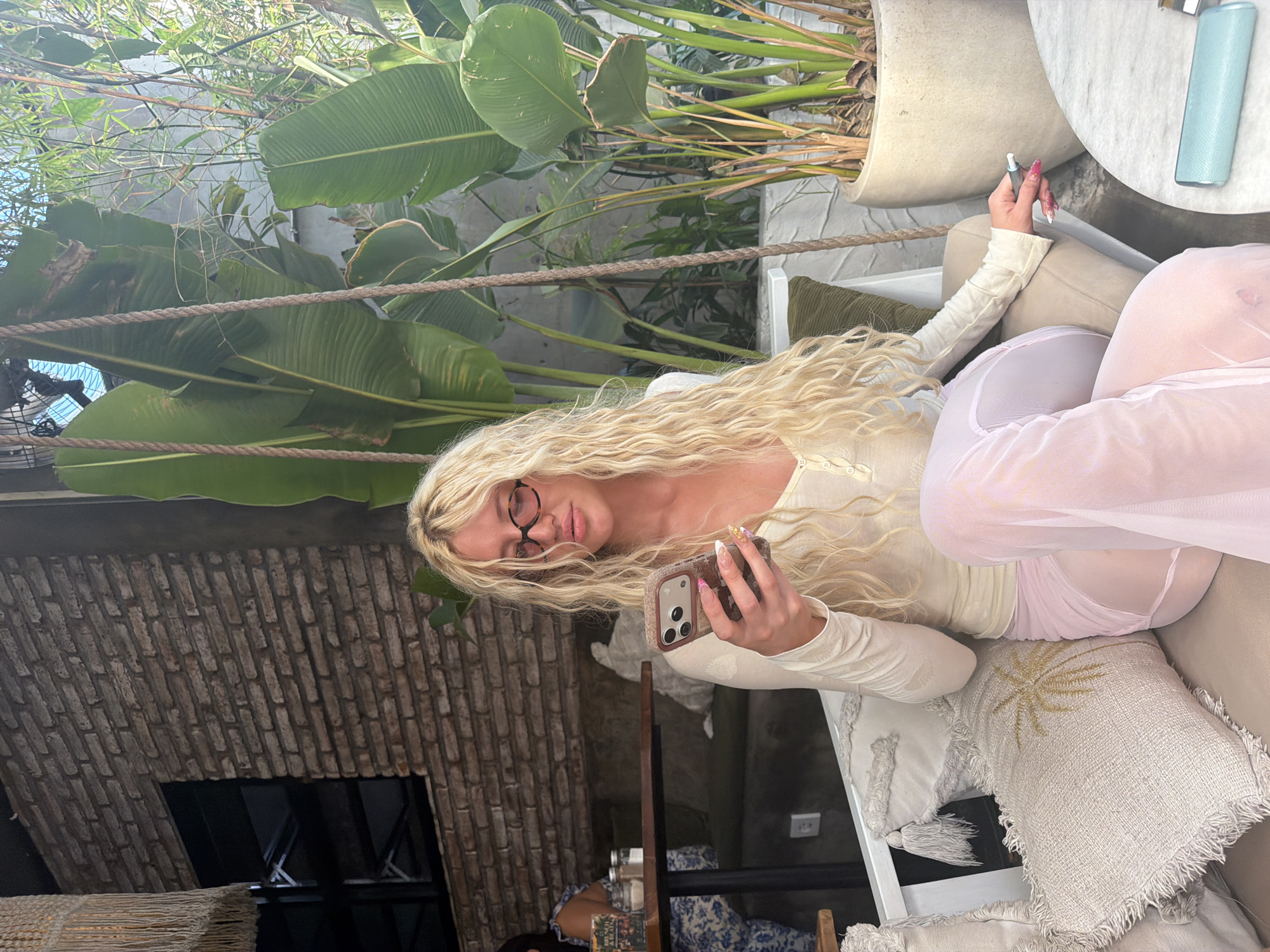 A woman with long blonde curly hair is seated on a swing indoors, looking at her phone. She is wearing a cream-colored long-sleeved shirt and sheer pink pants. She has on glasses and her nails are painted with bright colors. She is holding a vape pen in her right hand. The swing is suspended by thick ropes and has a beige seat. In front of her, there are several decorative pillows with tassels and a palm tree embroidery. Behind her, there is a brick wall and large tropical plants. To her right, there is a white circular table with a blue cylindrical object on it. In the background, another person is visible sitting at a table. The scene appears to be a cafe or restaurant with an indoor garden setting in North Kuta, Indonesia.