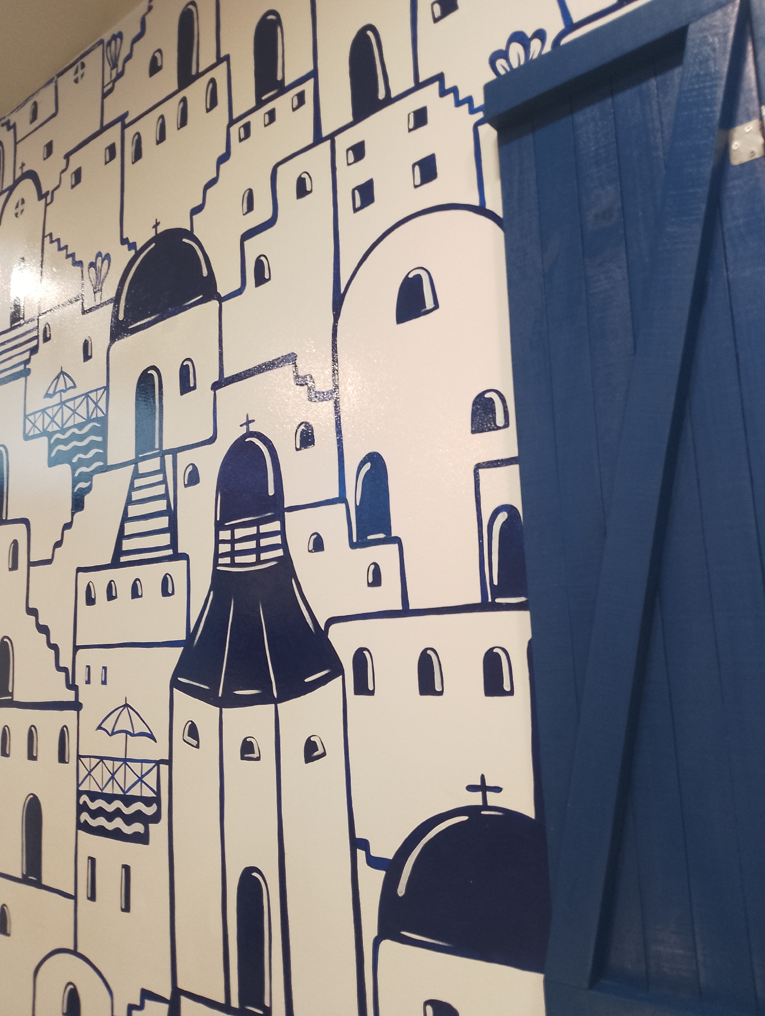 The image depicts a wall mural depicting a stylized Greek island town, rendered in white with blue line drawings. The scene includes depictions of buildings, churches, and architectural details suggestive of Santorini or Mykonos, complete with domes, archways, stairs, and coastal elements like a balcony with an umbrella. In the right foreground, a blue wooden shutter partially covers the mural. The scene does not show any people, actions or interactions. The location in the city of Pampatar, Venezuela is not shown or depicted in the image.