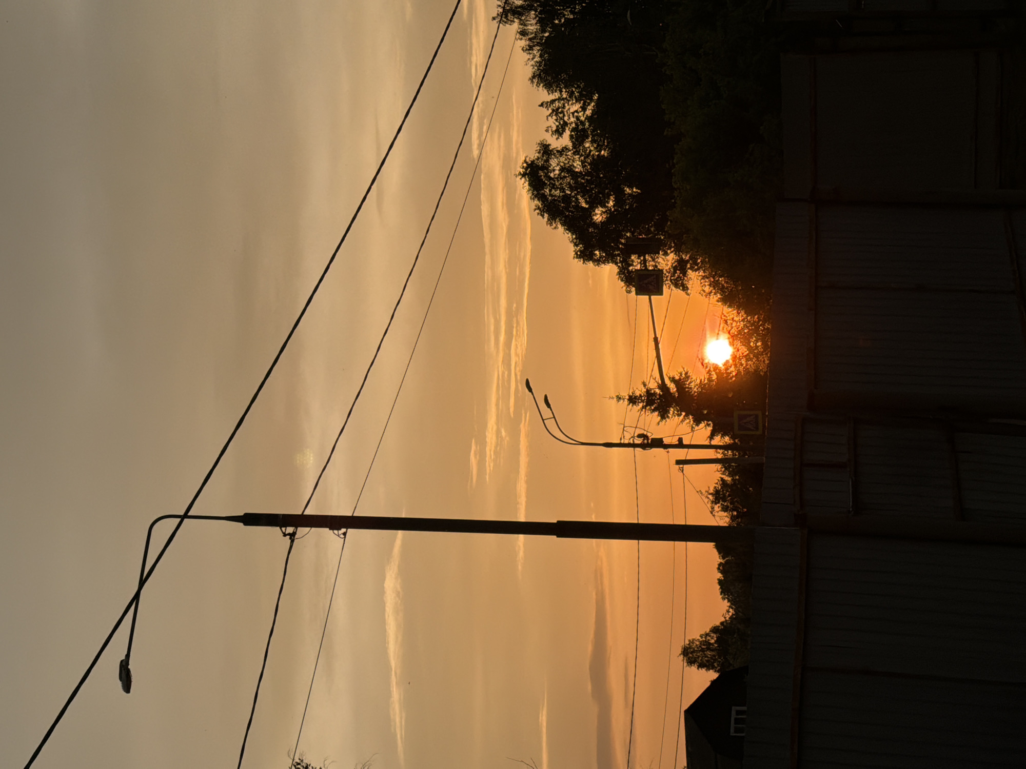 The image captures a sunset in the urban landscape of Спас-Заулок, Russia. In the foreground, a utility pole with attached power lines stretches vertically. Adjacent to the pole is a street lamp. Midground features trees and a pedestrian crossing sign silhouetted against the sun. The backdrop is an orange sky. To the right, a fence and part of a house are visible.
