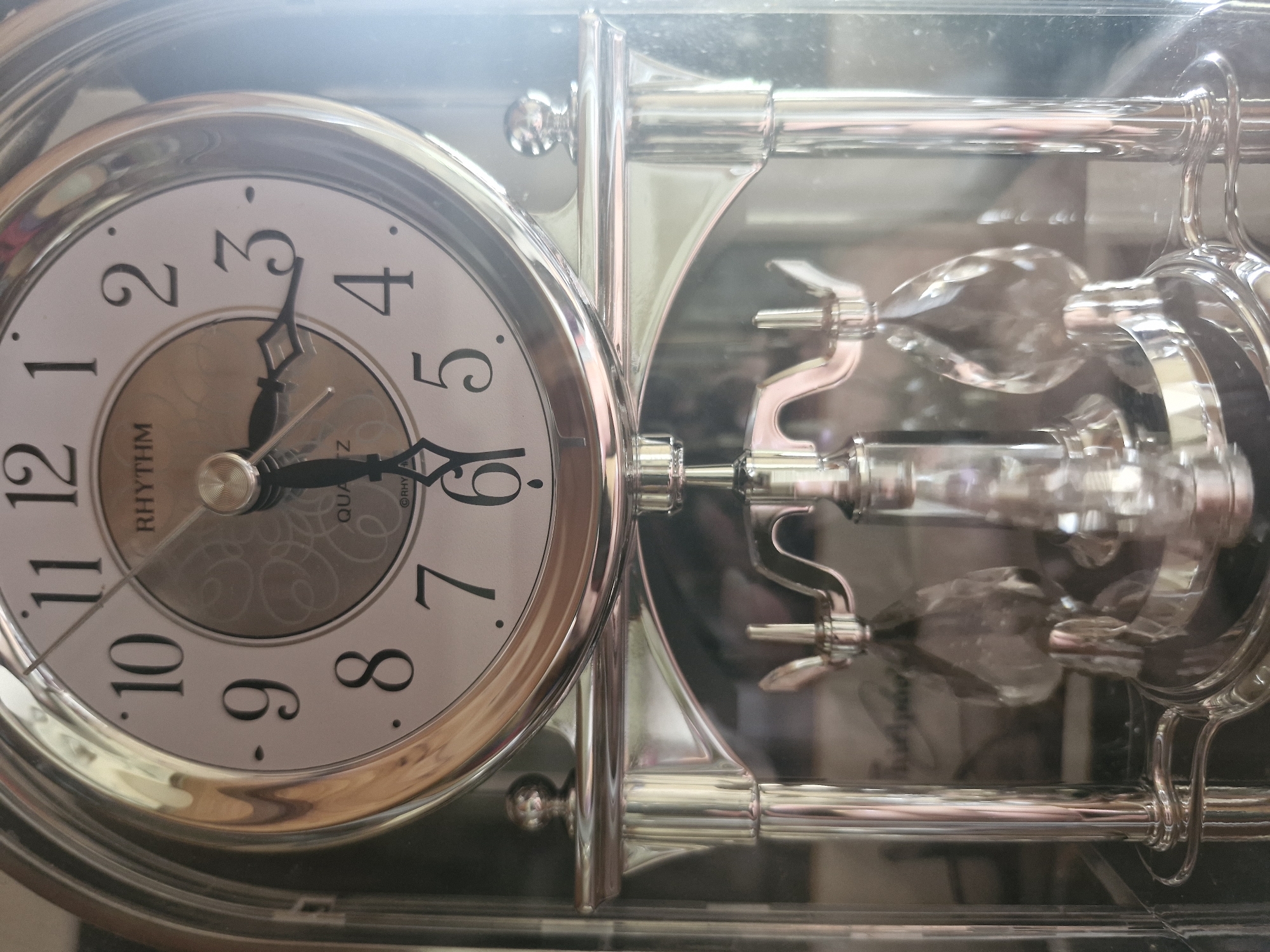A close-up shot of a clock face shows the time as approximately 10:17. The clock has a white face with black Roman numerals and hands. The clock is housed in a decorative, ornate silver-toned case with a pendulum visible at the bottom. The background is out of focus and appears to be a domestic interior. The image does not provide any information about the city of Ķekava, Latvia.