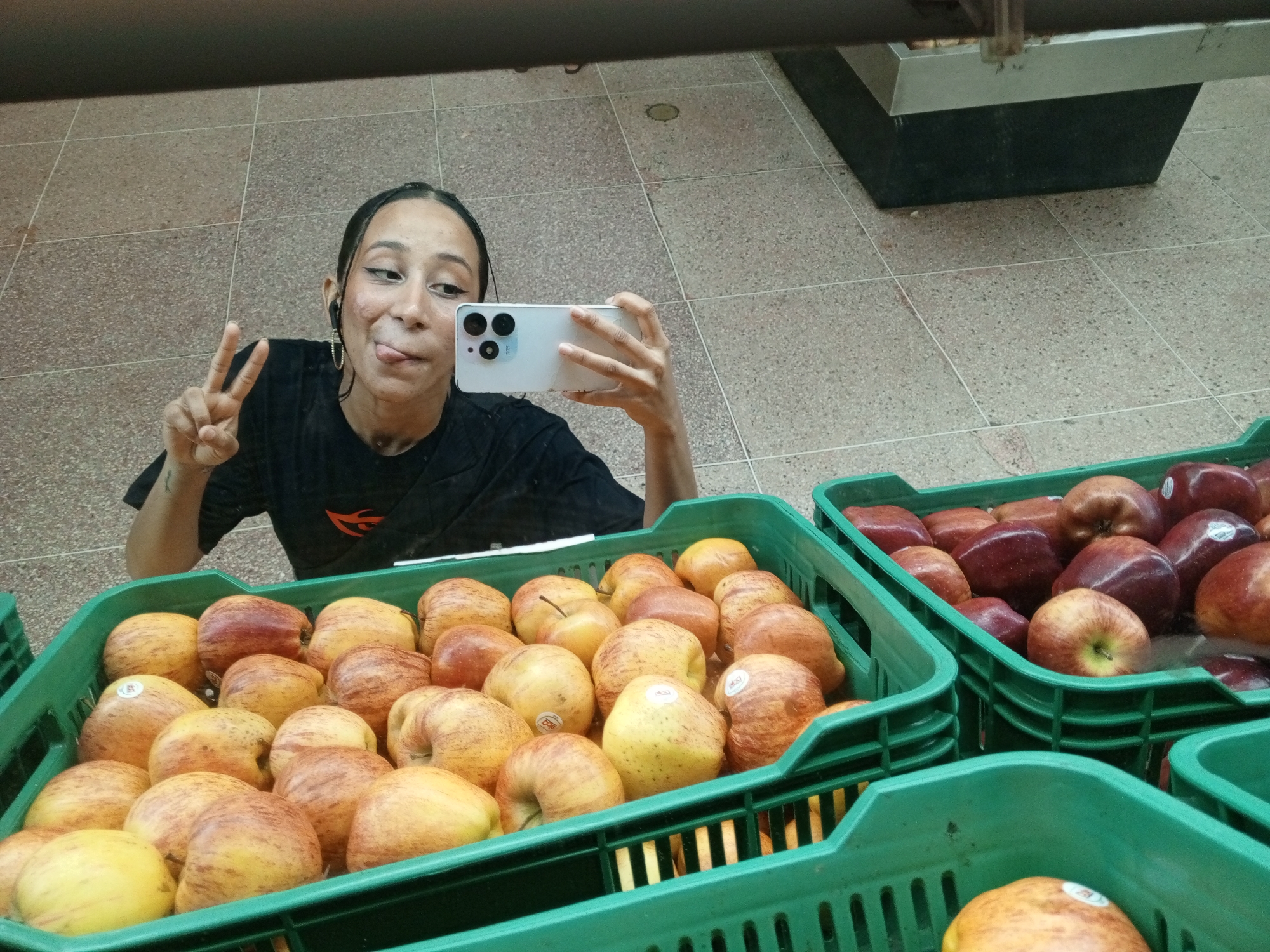A young adult woman takes a selfie in front of bins of apples, possibly at a supermarket in Porlamar, Venezuela. The woman has long black hair, visible tattoo on her left arm, and is wearing a black t-shirt and an earbud in her right ear. She holds a white smartphone in her right hand, and sticks out her tongue while holding up a "peace" sign with her left hand. In the foreground, there are three green plastic bins filled with apples. Two bins appear to contain yellow-red apples and the other bin has red apples. The floor is tiled and there is what appears to be a stainless steel refrigeration unit in the background. The image seems to be taken in front of a mirror.